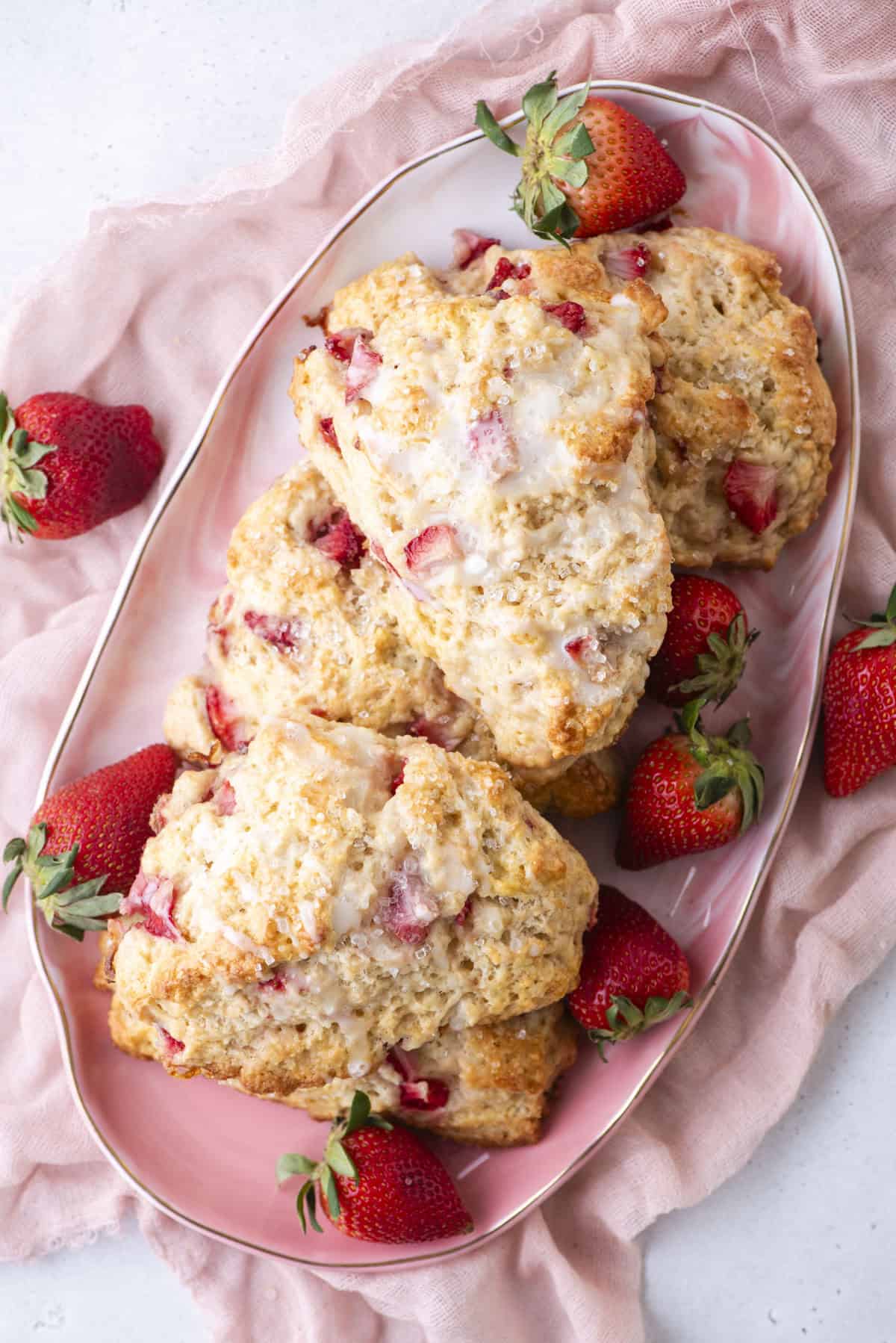 A plate of strawberry scones topped with icing, surrounded by fresh strawberries on a pink cloth. The scones are golden brown with visible strawberry pieces, creating a delicious and inviting presentation.