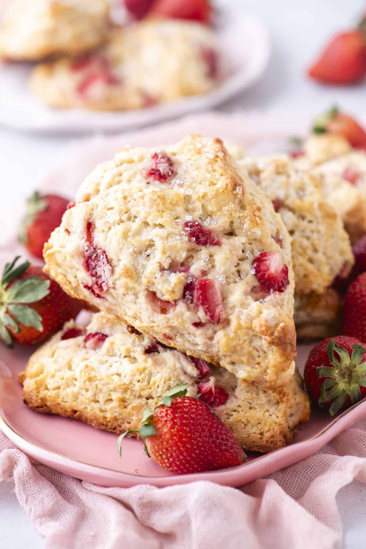 Close-up of two scones studded with fresh strawberries, stacked on a pink plate. More strawberries and additional scones are in the background. The scones are golden-brown with a flaky texture. A soft pink cloth partially covers the surface.