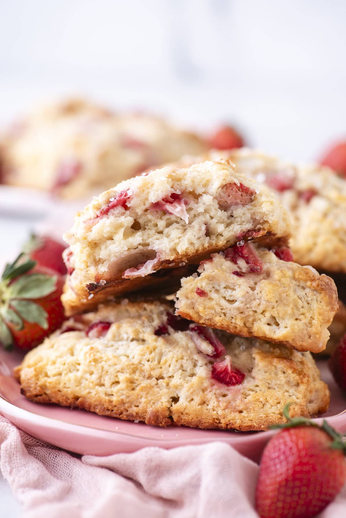 Close-up of freshly baked scones filled with sliced strawberries, stacked on a pink plate. Whole strawberries surround the plate, and a soft pink cloth is partially visible in the foreground.