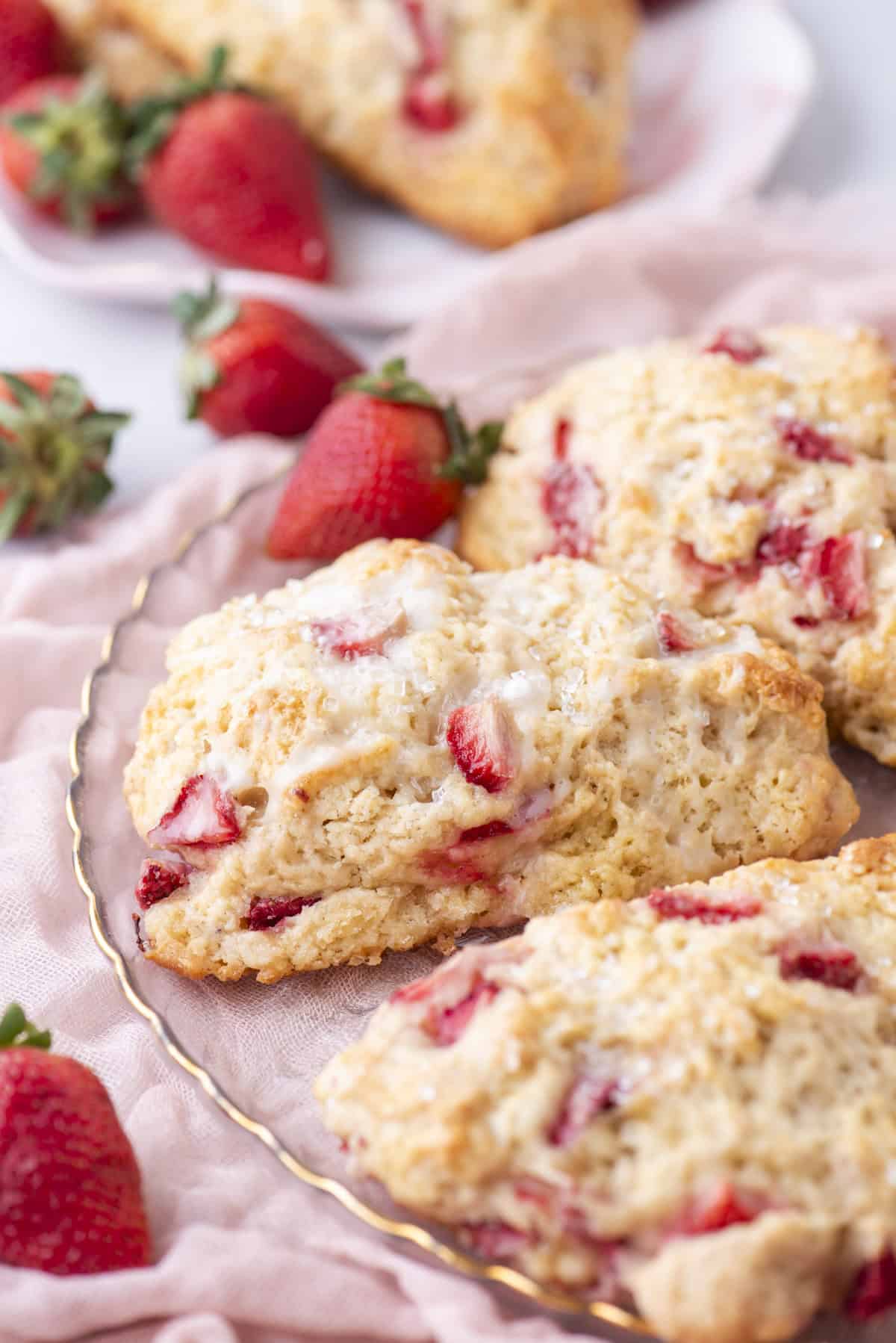 Close-up of freshly baked strawberry scones on a decorative plate. The scones are golden brown with visible strawberry pieces and a light glaze on top. Fresh strawberries are scattered around, and a soft pink cloth is partially visible underneath the plate.