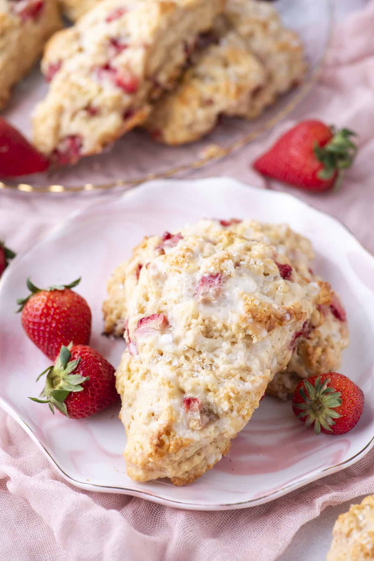 A plate with a strawberry scone garnished with fresh strawberries. The scone has a golden, crumbly texture and visible strawberry pieces. More scones and strawberries are in the background, set on a soft pink fabric.