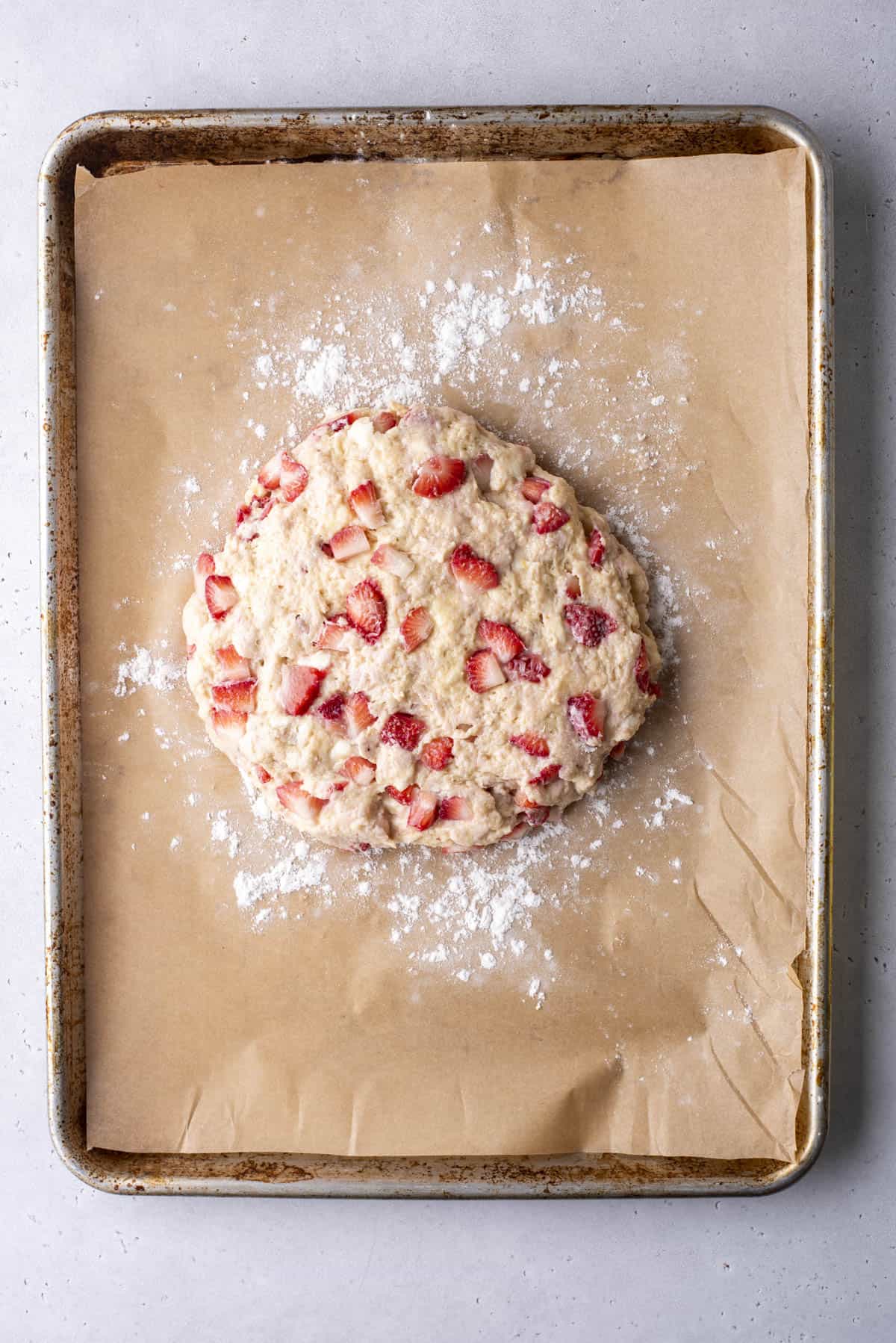 Round scone dough with strawberries on parchment paper-lined baking sheet, dusted with flour.