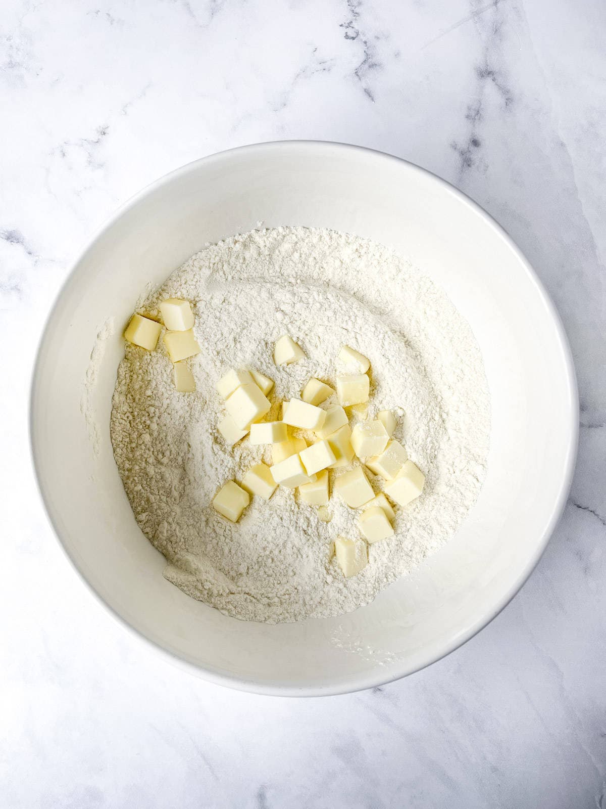 A white bowl containing flour with small cubes of butter on top. The bowl is placed on a marble countertop.