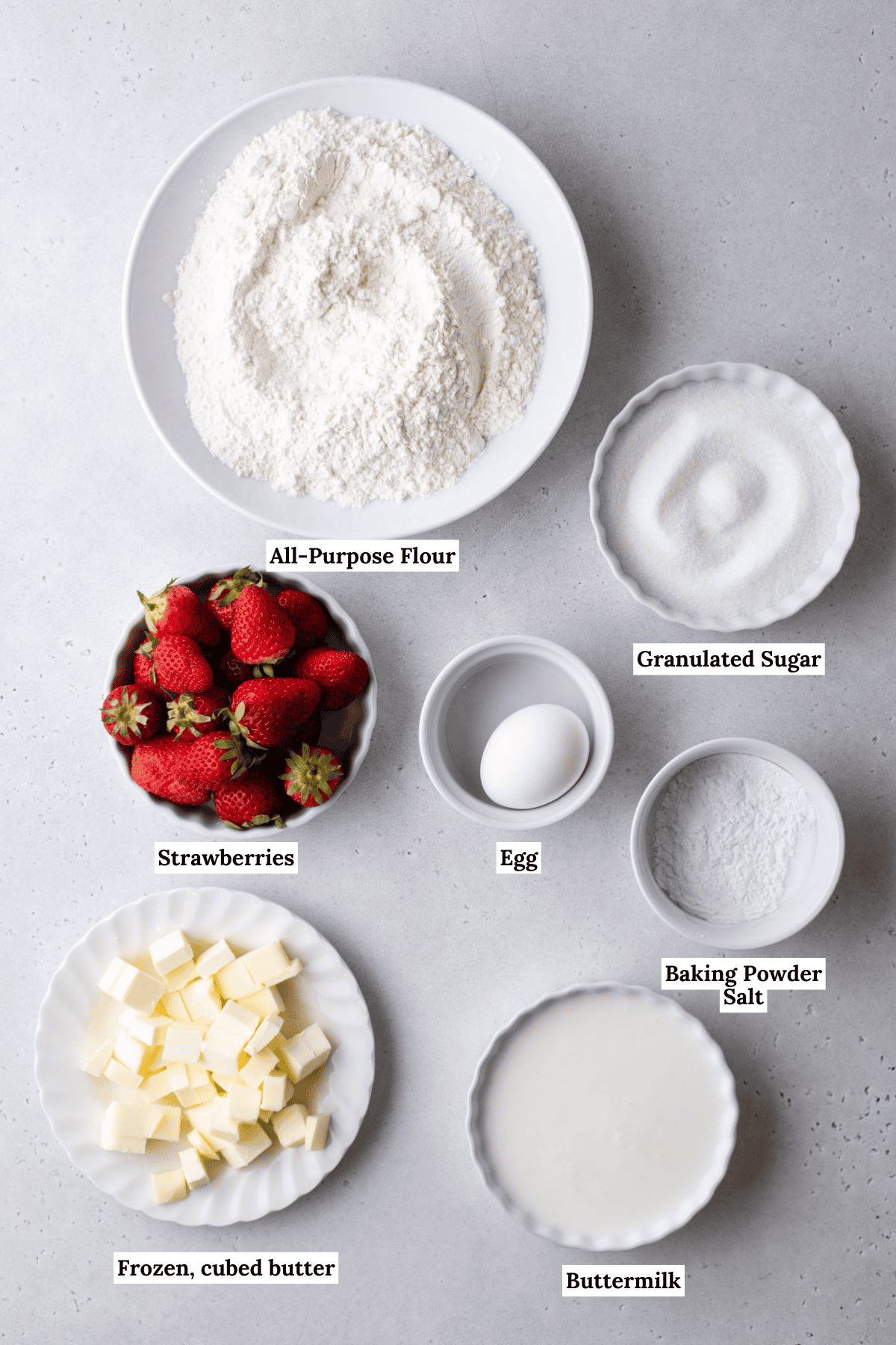 A top view of baking ingredients on a gray surface, including bowls of all-purpose flour, granulated sugar, baking powder, salt, an egg, cubed frozen butter, buttermilk, and fresh strawberries.