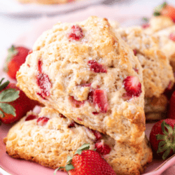 A plate of strawberry scones, golden-brown and studded with red strawberry pieces, sits on a pink plate. Fresh strawberries are scattered around. A red banner above reads Strawberry Scones.