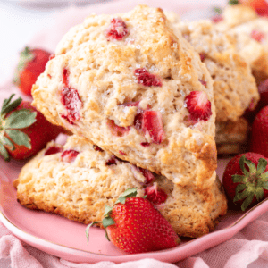 Close-up of two strawberry scones topped with sugar crystals on a pink plate. Fresh strawberries are scattered around the scones, and a light pink cloth is in the background, creating a soft, inviting atmosphere.