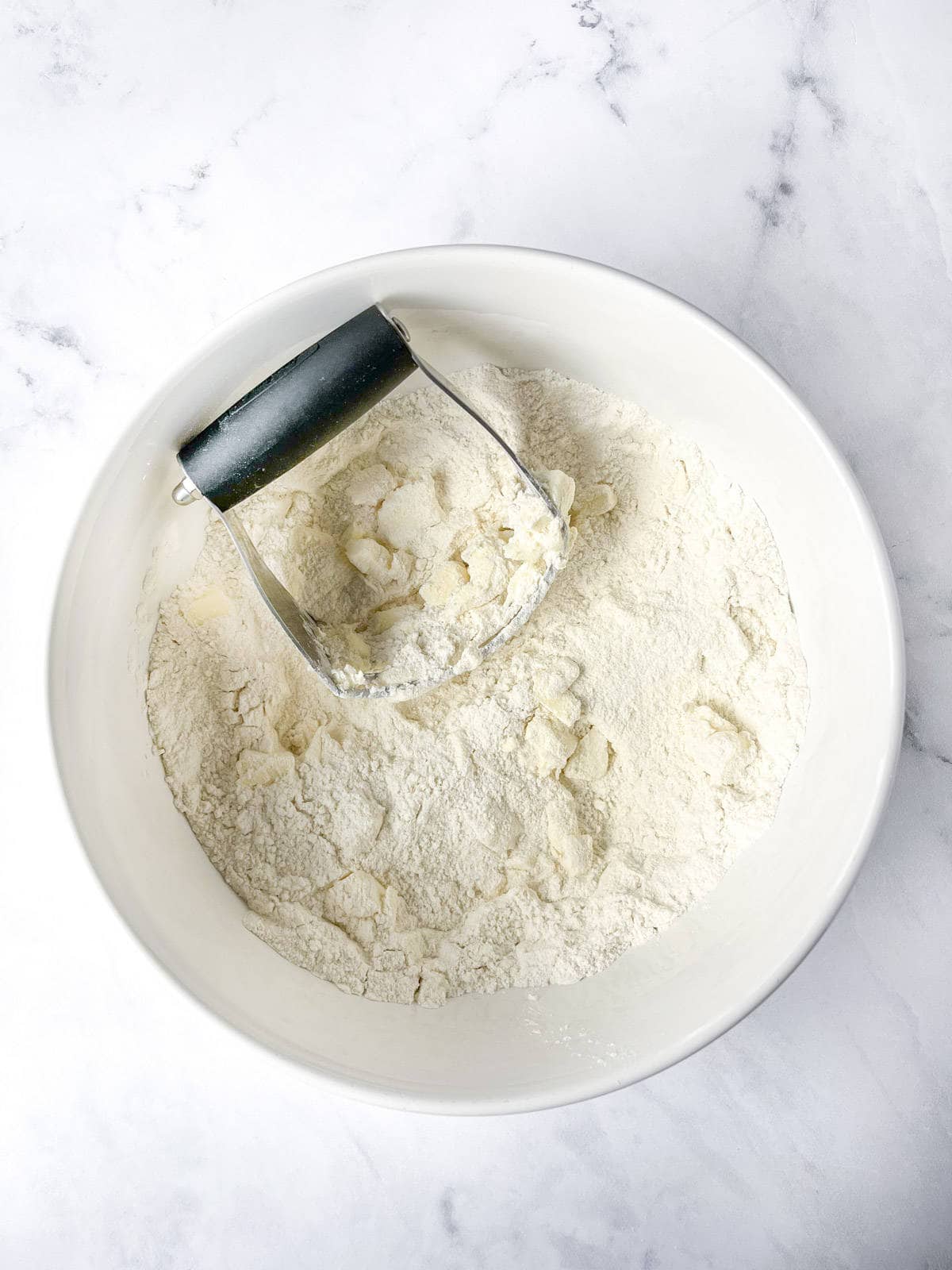 A white bowl on a marble surface contains a mixture of flour and butter chunks. A pastry cutter with a black handle is partially submerged in the mixture, indicating the process of cutting butter into the flour.