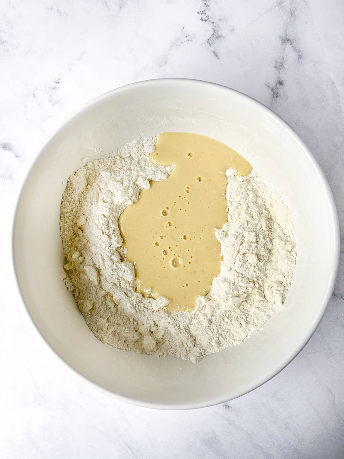 A white bowl containing flour with a pool of liquid in the center, ready for mixing. The bowl is placed on a marble surface.