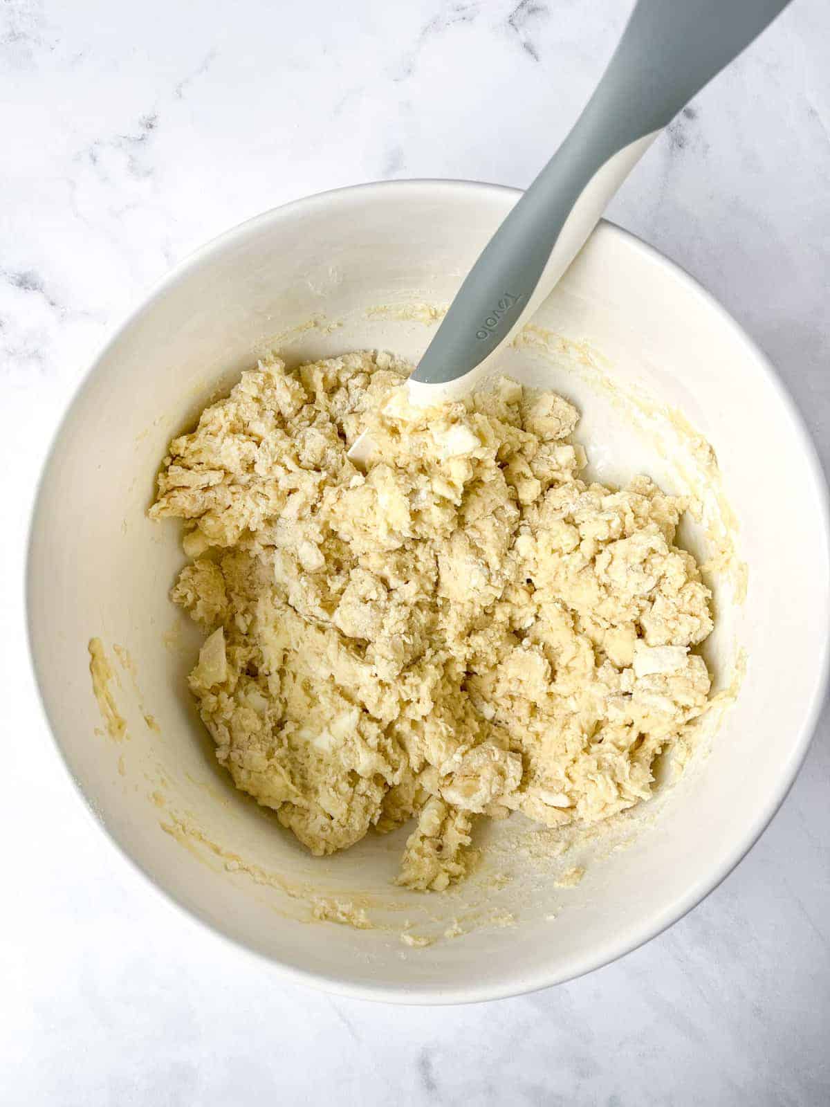 A white mixing bowl containing crumbly biscuit dough with chunks of butter is being stirred by a gray silicone spatula. The bowl rests on a marbled countertop.