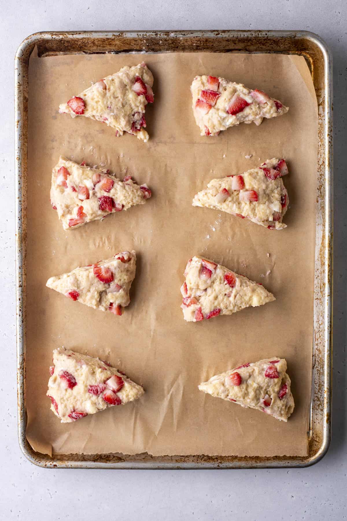 Eight unbaked strawberry scones are arranged on a parchment-lined baking sheet. The scones are triangular with visible strawberry chunks, ready for baking.