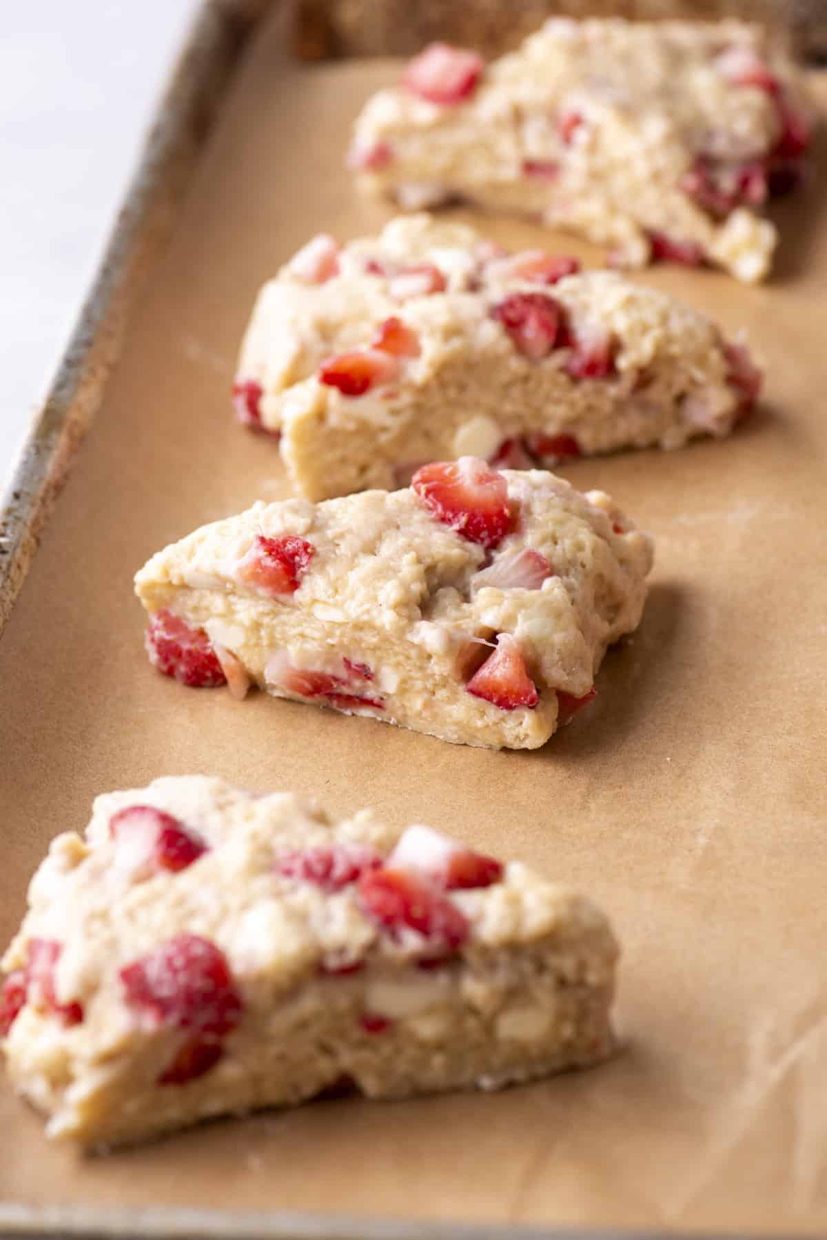 Unbaked strawberry scones arranged on a parchment-lined baking sheet. The scones are triangular and filled with visible strawberry chunks, ready to be baked.