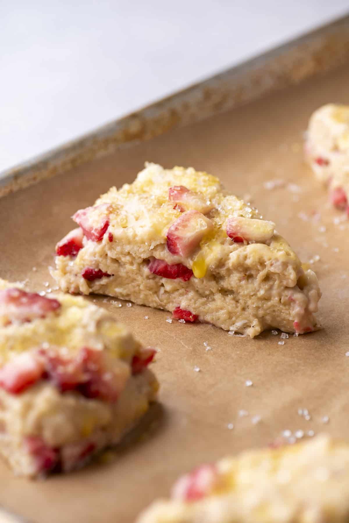 Close-up of a triangular strawberry scone on a parchment-lined baking sheet. The scone is golden-brown with visible chunks of strawberries and a sprinkle of sugar on top. Other scones are partially visible in the background.