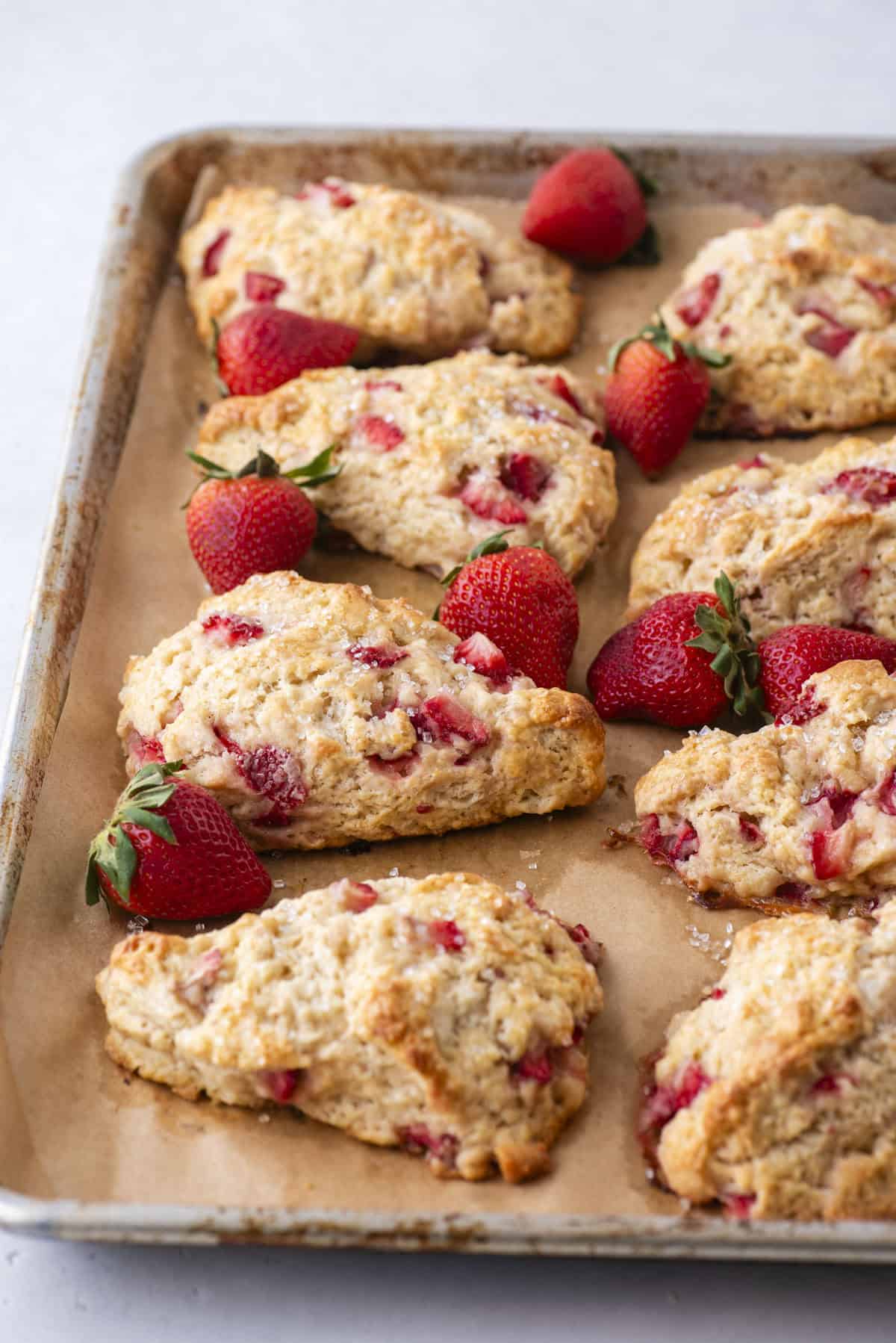 A baking tray with parchment paper holds several freshly baked scones filled with pieces of strawberries. Whole strawberries are scattered around the scones, adding color to the scene.