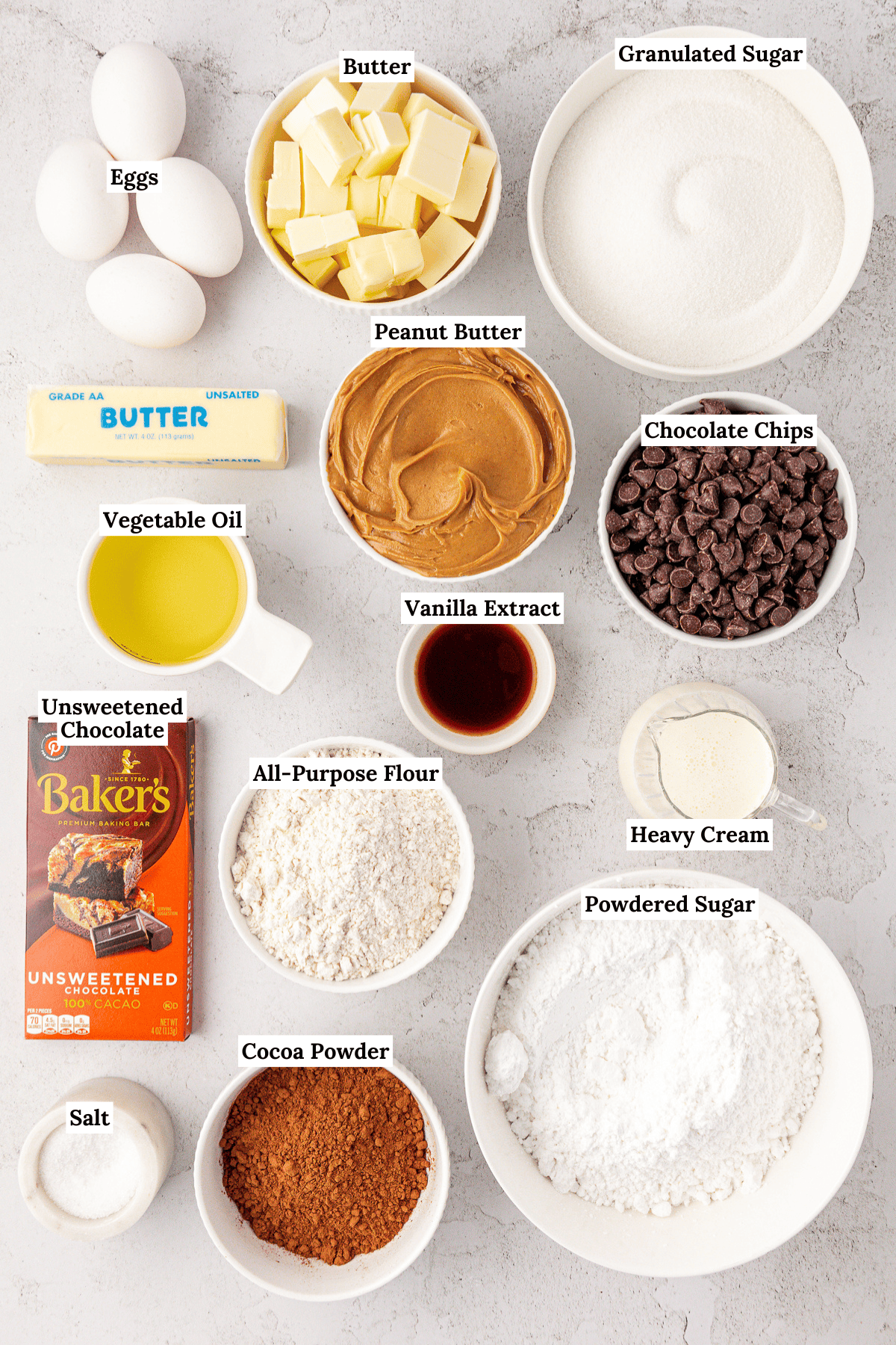 A variety of baking ingredients are arranged in bowls and on the counter, including eggs, butter, granulated sugar, peanut butter, chocolate chips, oil, flour, heavy cream, unsweetened chocolate, cocoa powder, powdered sugar, vanilla, and salt.