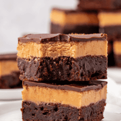 Two thick, square buckeye brownies on a white plate, featuring a dark chocolate brownie base, a creamy peanut butter layer, and glossy chocolate ganache on top. Other brownies are blurred in the background.
