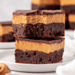 Two decadent brownies stacked on a white plate, each with a fudgy chocolate base, a thick creamy peanut butter layer, and a glossy chocolate topping. Other brownies are blurred in the background.