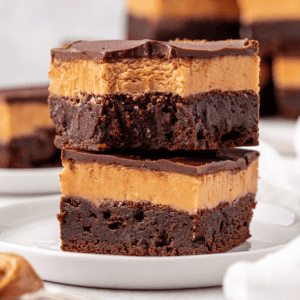 Two decadent brownies stacked on a white plate, each with a fudgy chocolate base, a thick creamy peanut butter layer, and a glossy chocolate topping. Other brownies are blurred in the background.