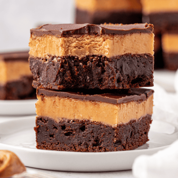 Two decadent brownies stacked on a white plate, each with a fudgy chocolate base, a thick creamy peanut butter layer, and a glossy chocolate topping. Other brownies are blurred in the background.