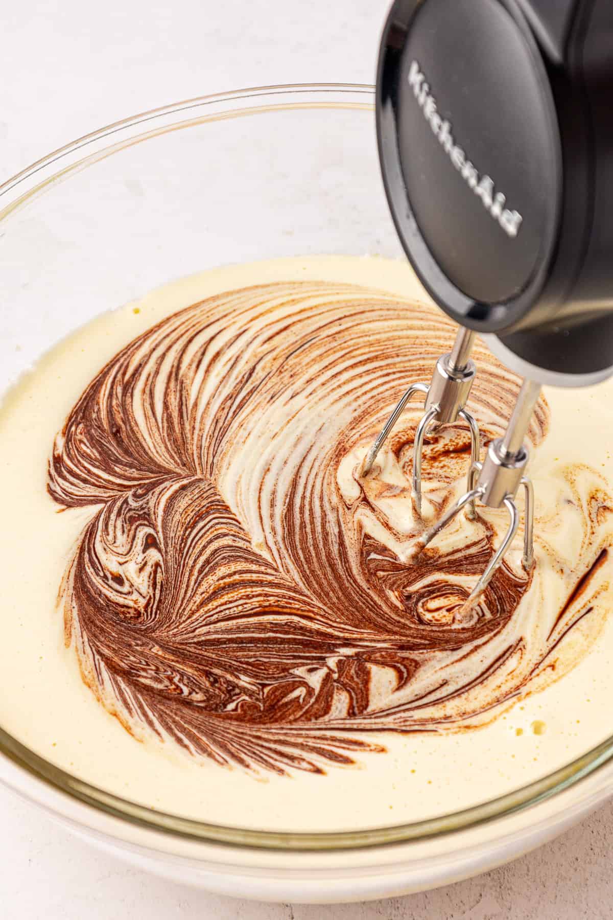 A stand mixer blends light yellow batter with swirls of chocolate in a clear glass bowl, creating a marbled effect.