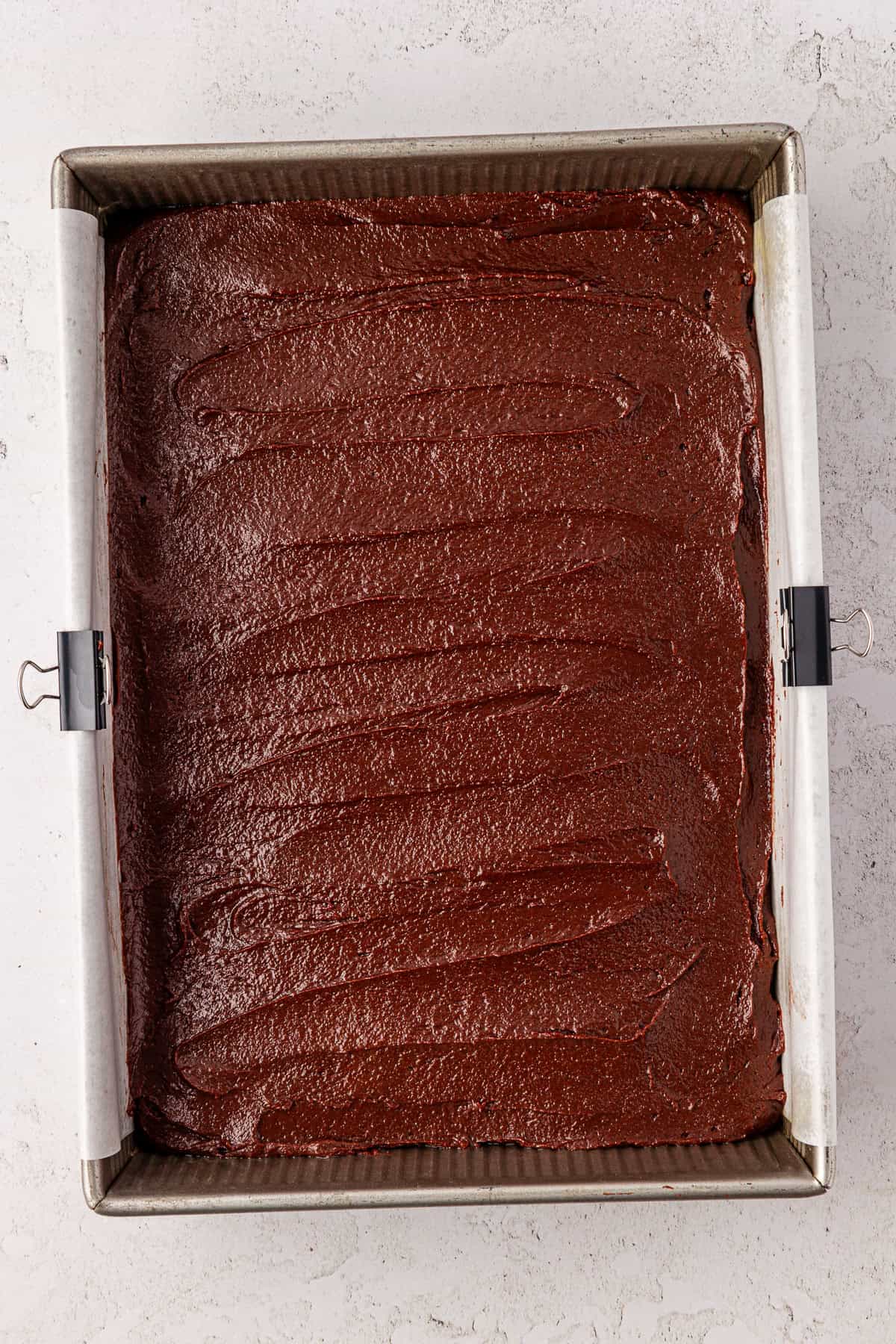 A square baking pan lined with parchment paper holds an even layer of chocolate brownie batter, ready to be baked. The batter is glossy and spread smoothly with visible swirls on the surface.