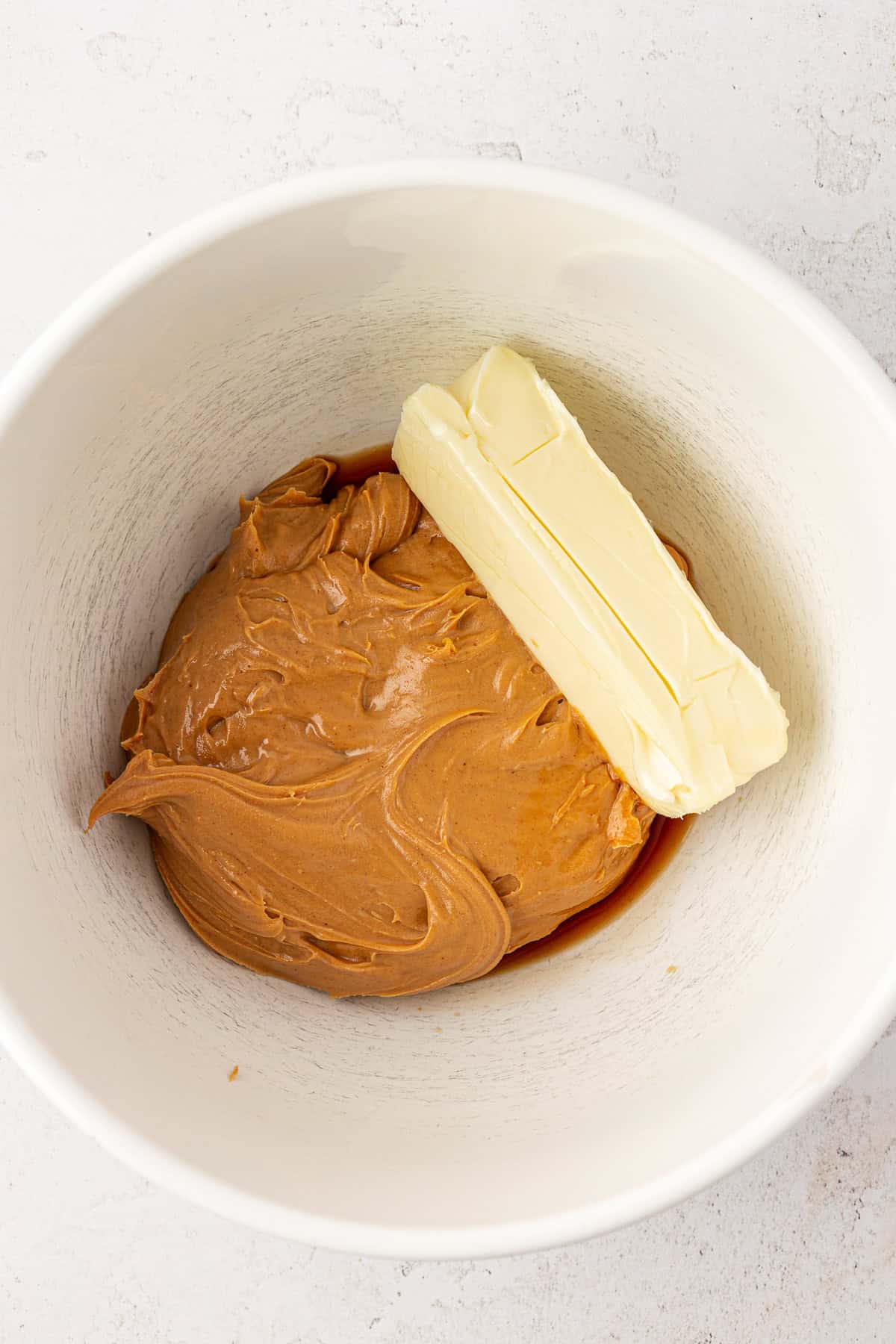 A white mixing bowl containing a large dollop of peanut butter and a stick of butter on a light-colored surface.