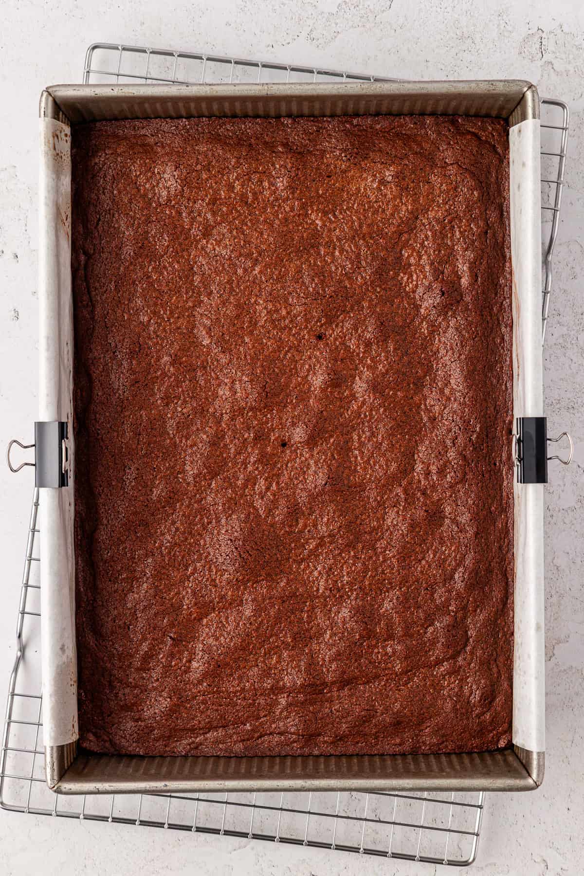 A square pan of freshly baked brownies, lined with parchment paper and clipped at the sides, sits on a metal cooling rack atop a white surface.
