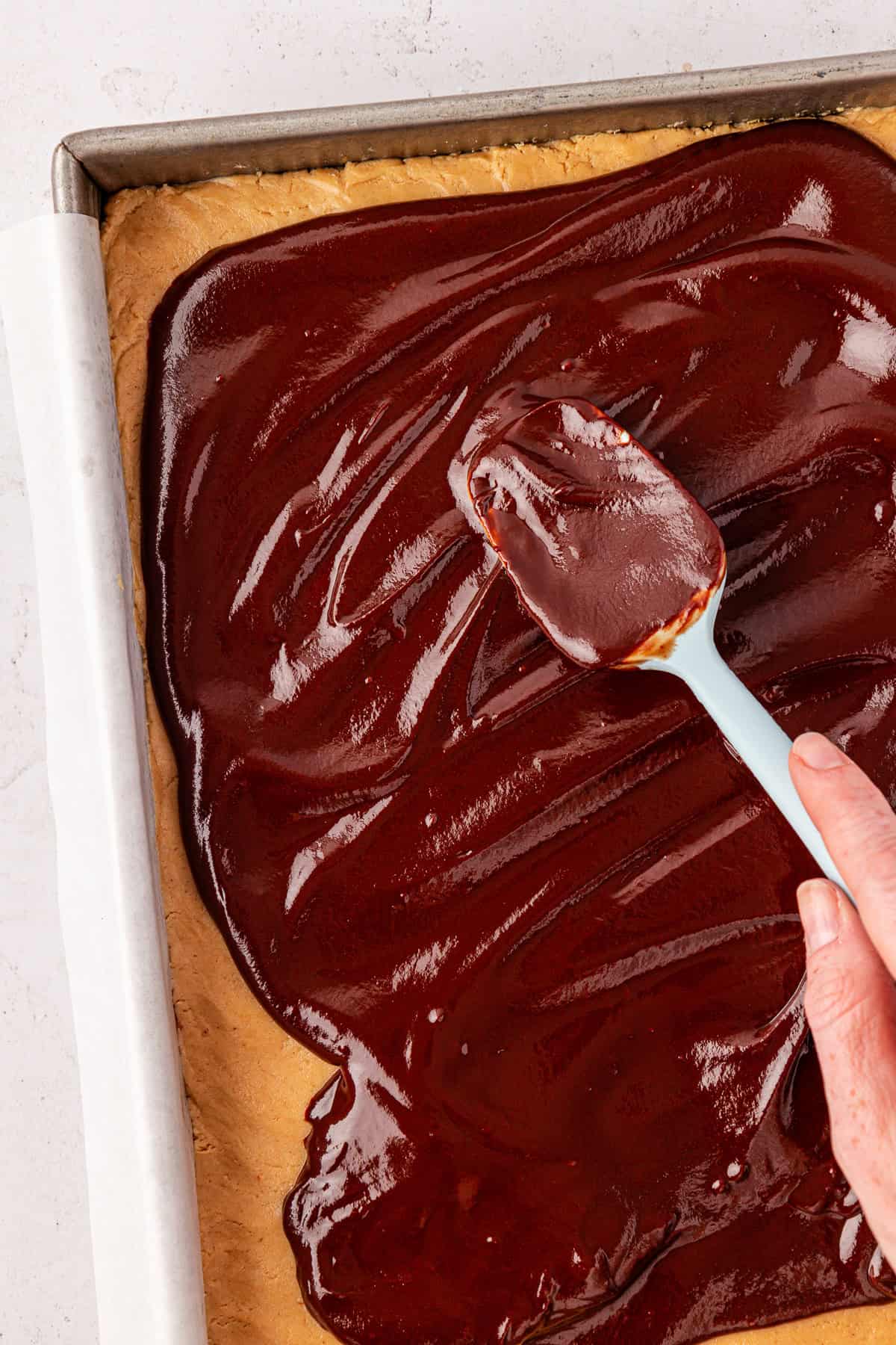 A hand spreads glossy chocolate frosting over a rectangular sheet of buckeye brownies in a parchment-lined baking pan.