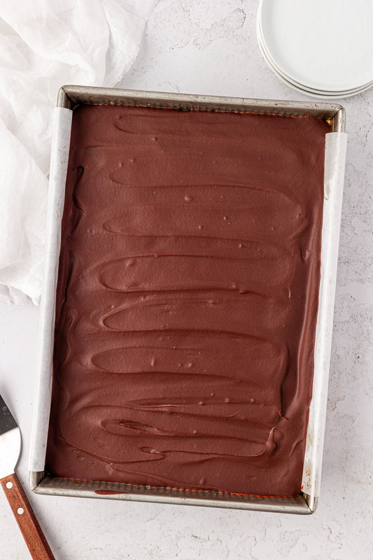 A rectangular baking pan filled with buckeye brownies, set on a light textured surface with a white cloth, round plates, and a cake server nearby.