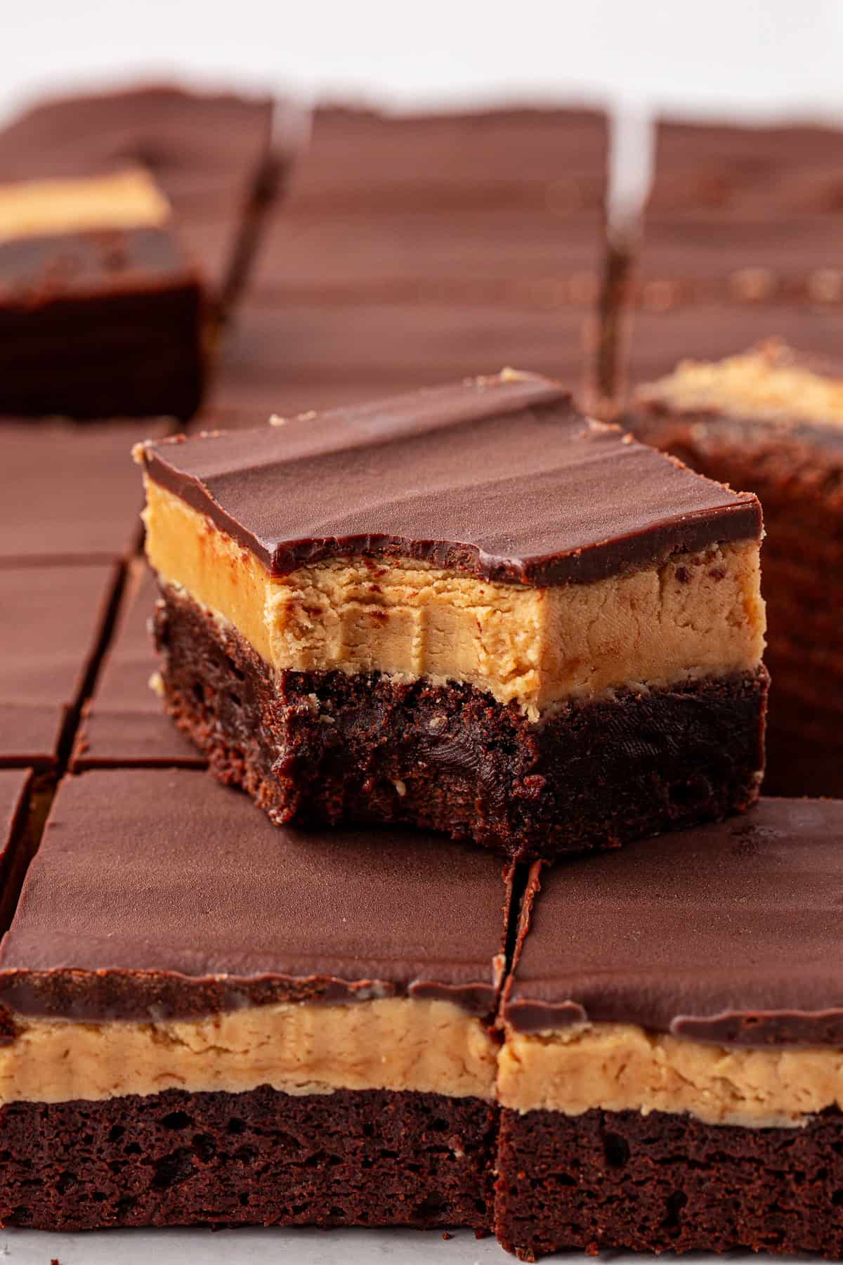 A close-up of chocolate and peanut butter layered buckeye brownies, with one brownie on top showing a bite taken out, revealing the creamy peanut butter and rich chocolate layers.