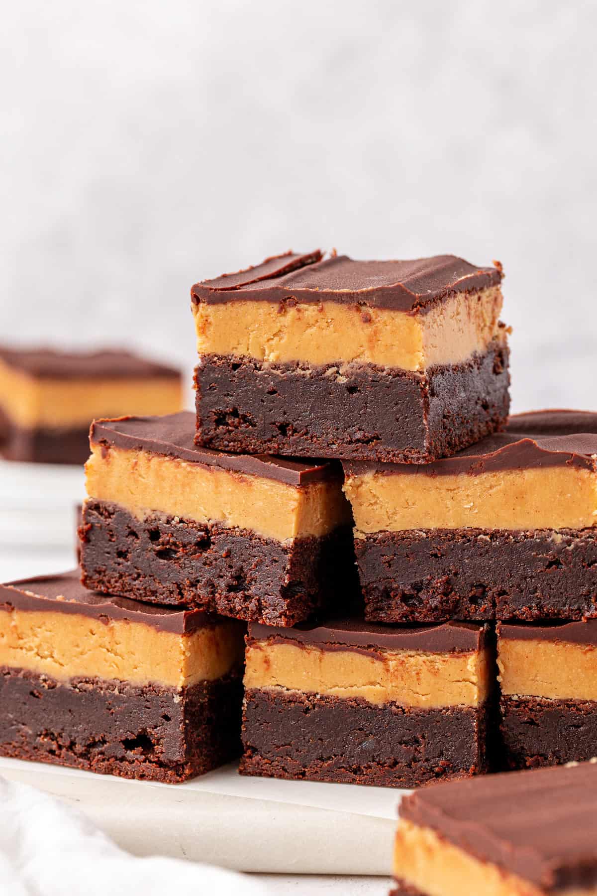 A stack of rich buckeye brownies with a thick peanut butter layer and a smooth chocolate topping, arranged on a white surface with more brownies blurred in the background.