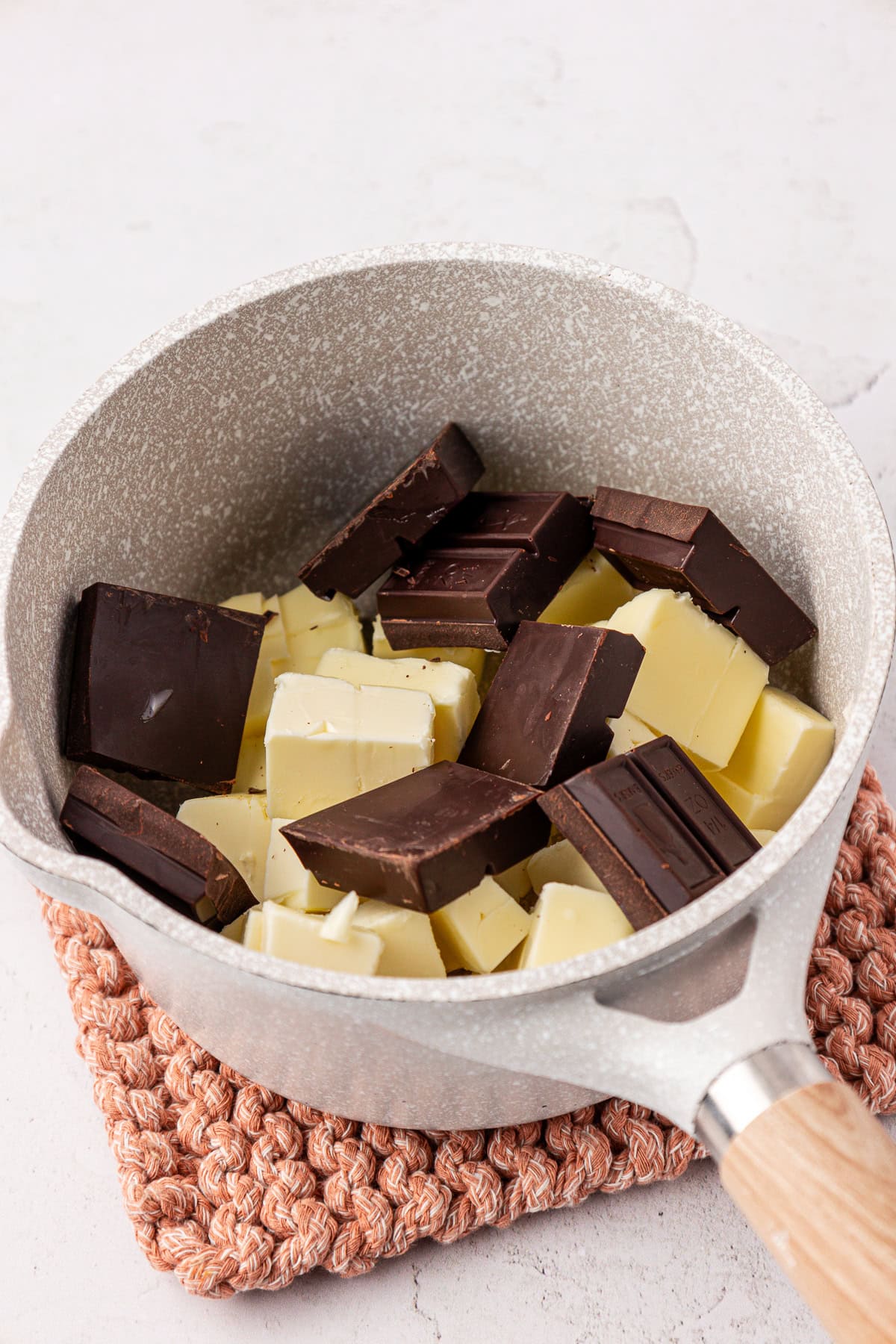 A saucepan containing chunks of white and dark chocolate sits on a textured pink trivet atop a light-colored surface.