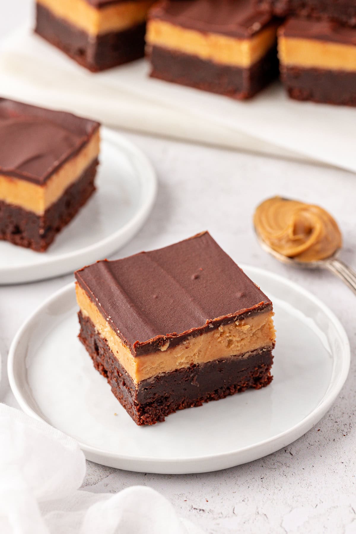 A close-up of a buckeye brownie with three layers: a dense brownie base, a creamy peanut butter middle, and a smooth chocolate topping, served on a white plate. A spoon with peanut butter and more dessert bars are in the background.