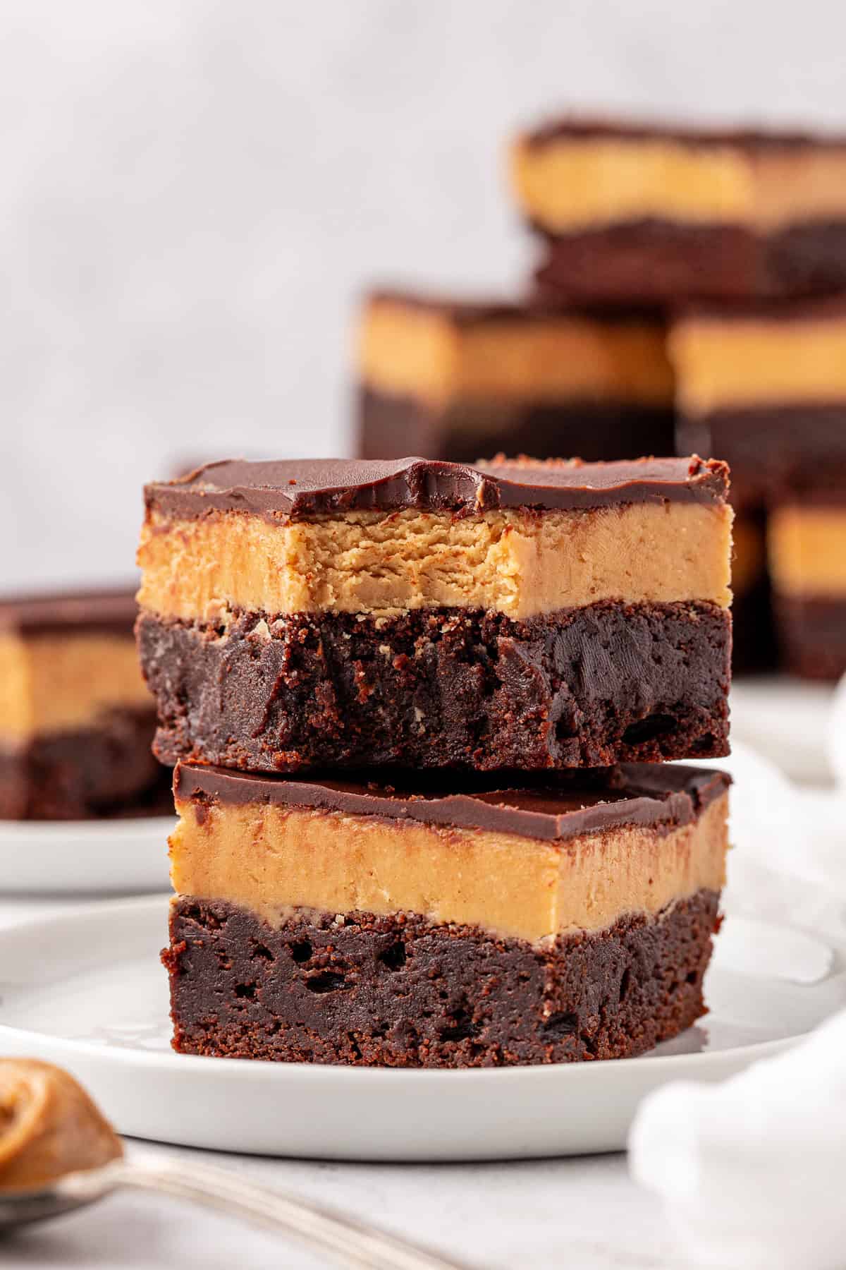 Two buckeye brownies with a thick peanut butter layer and chocolate topping are stacked on a white plate. More brownies are visible in the background, slightly out of focus.
