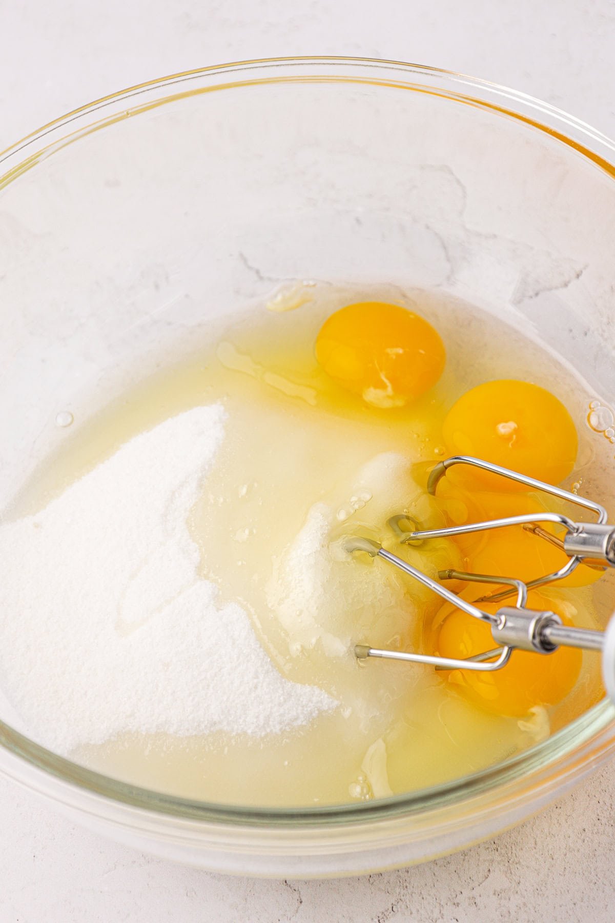 A glass bowl containing four raw eggs, sugar, and a hand mixer with metal beaters, ready for mixing on a light-colored countertop.