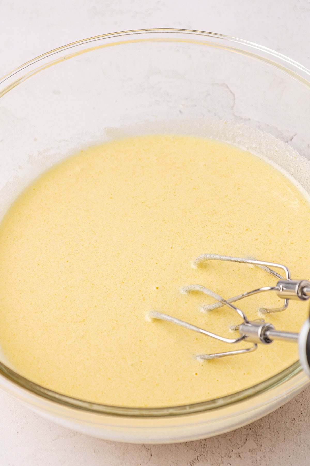 A close-up of a glass mixing bowl filled with yellow cake batter. Two metal beaters from a hand mixer are partially visible on the right side of the bowl. The bowl sits on a light-colored surface.