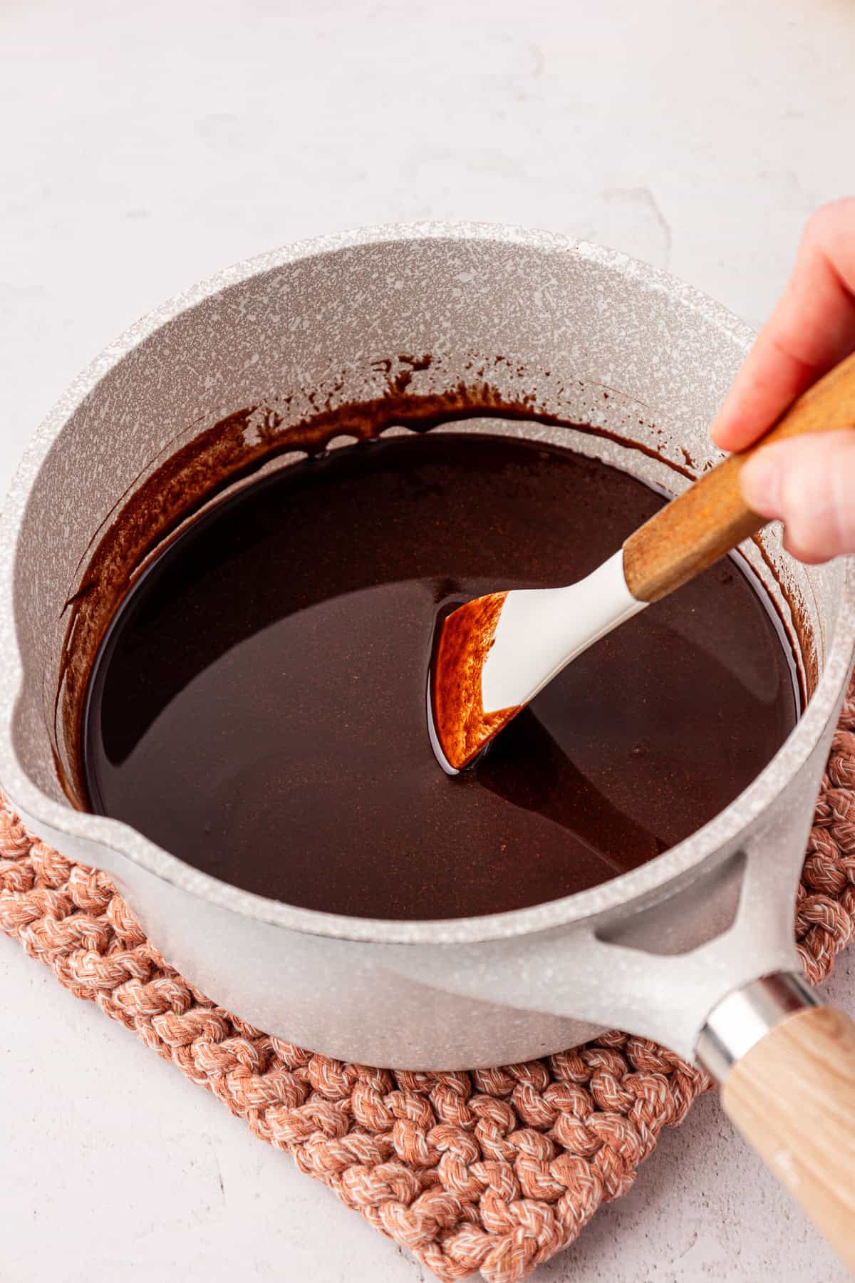 A hand stirs glossy melted chocolate in a white saucepan using a white and wooden spatula, with the pot placed on a pink textured trivet.