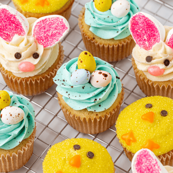 A variety of Easter-themed cupcakes on a wire rack, decorated as chicks with yellow icing, bunnies with pink ears, and some with blue swirled frosting topped with candy eggs.