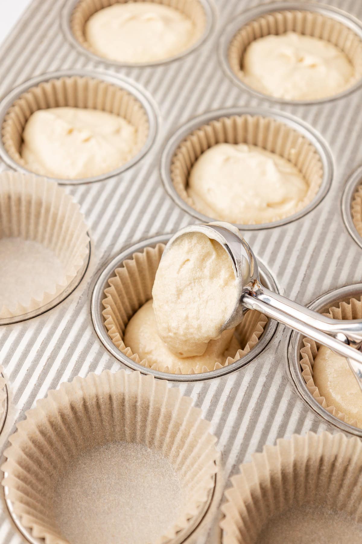 A metal scoop fills a paper-lined muffin tin with batter. Some cups are already filled, while others are empty and ready for batter to be added.