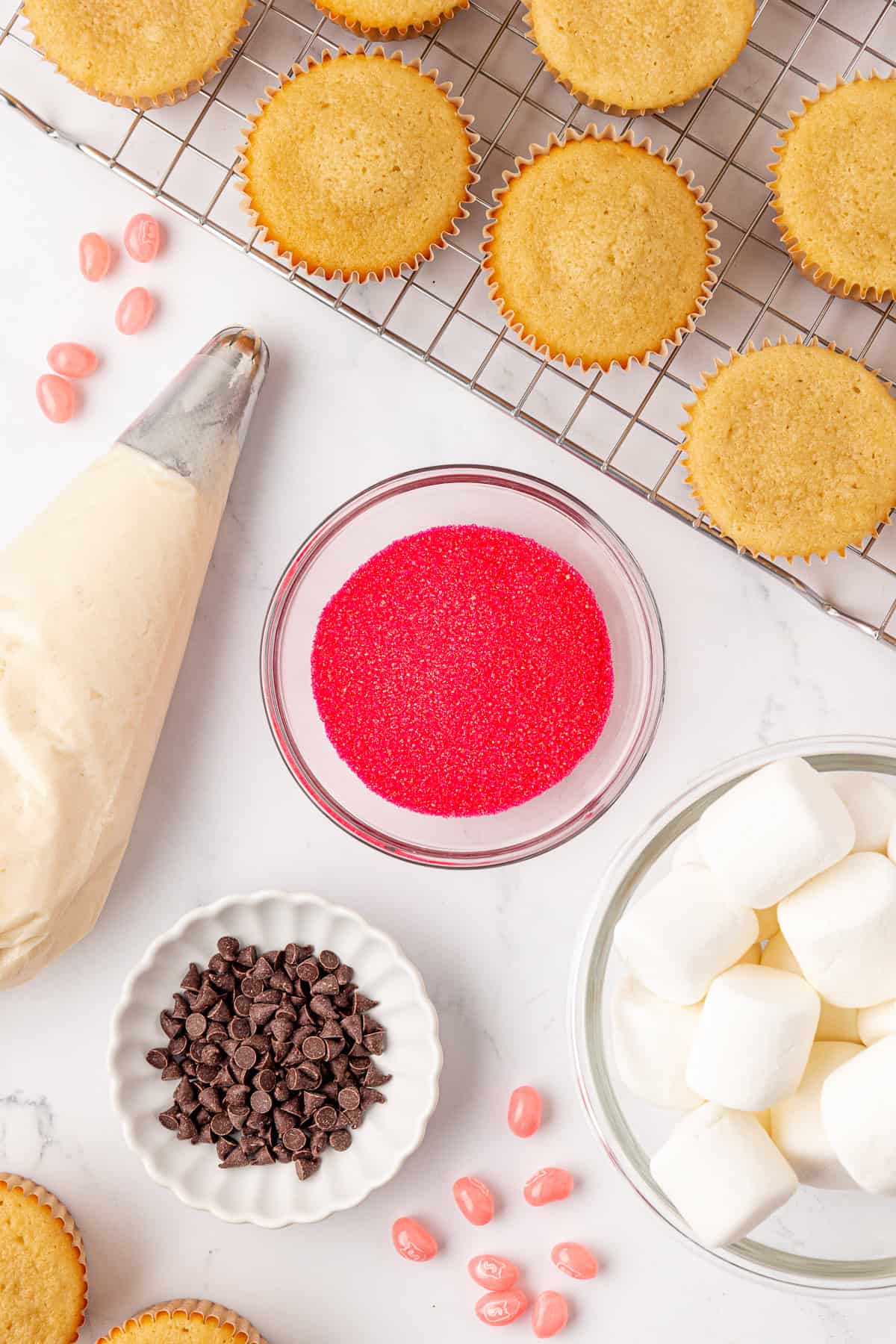 Cupcakes on a cooling rack, with baking supplies nearby: a piping bag filled with frosting, a bowl of red sugar sprinkles, chocolate chips, large marshmallows, and pink candy pieces on a white surface.