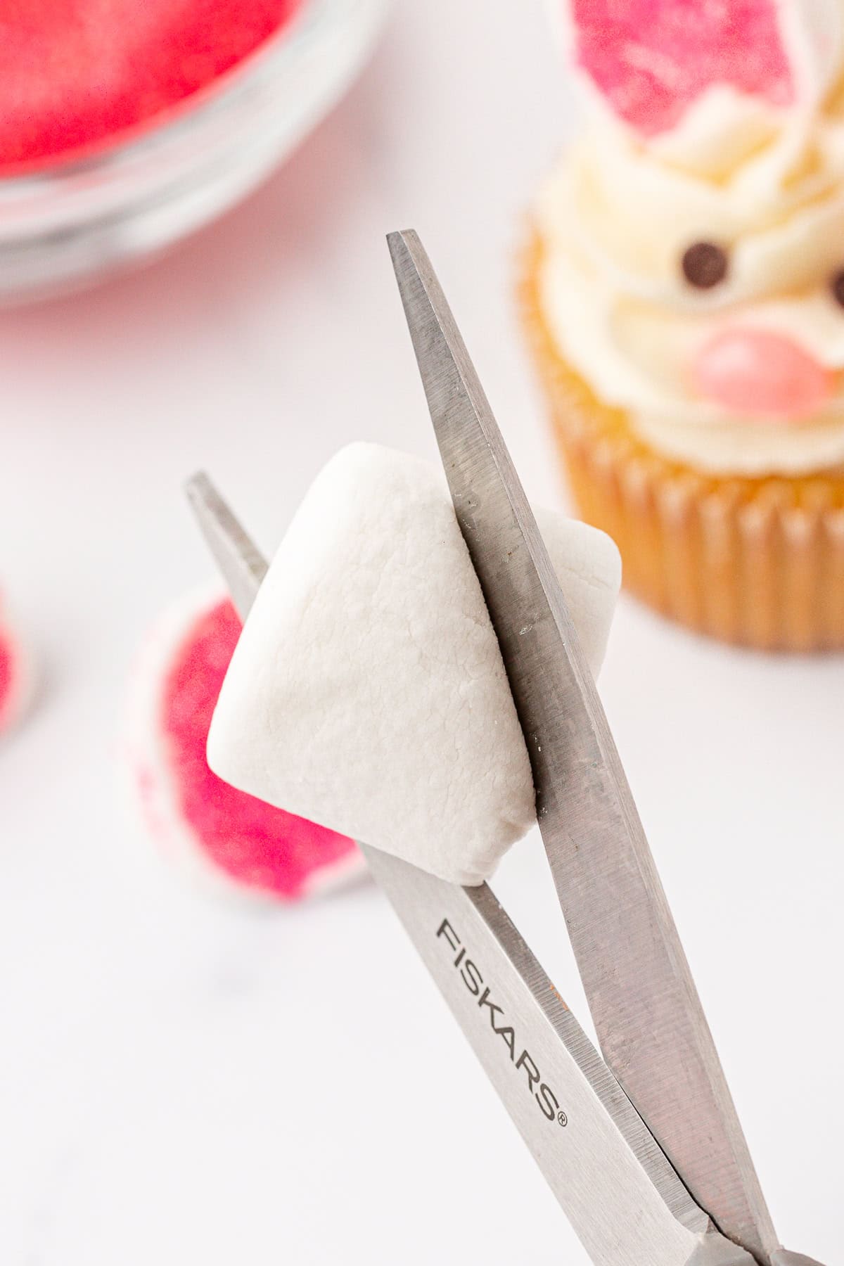 A pair of scissors cutting a white marshmallow, with a cupcake decorated like a bunny and a plate of pink treats in the background.