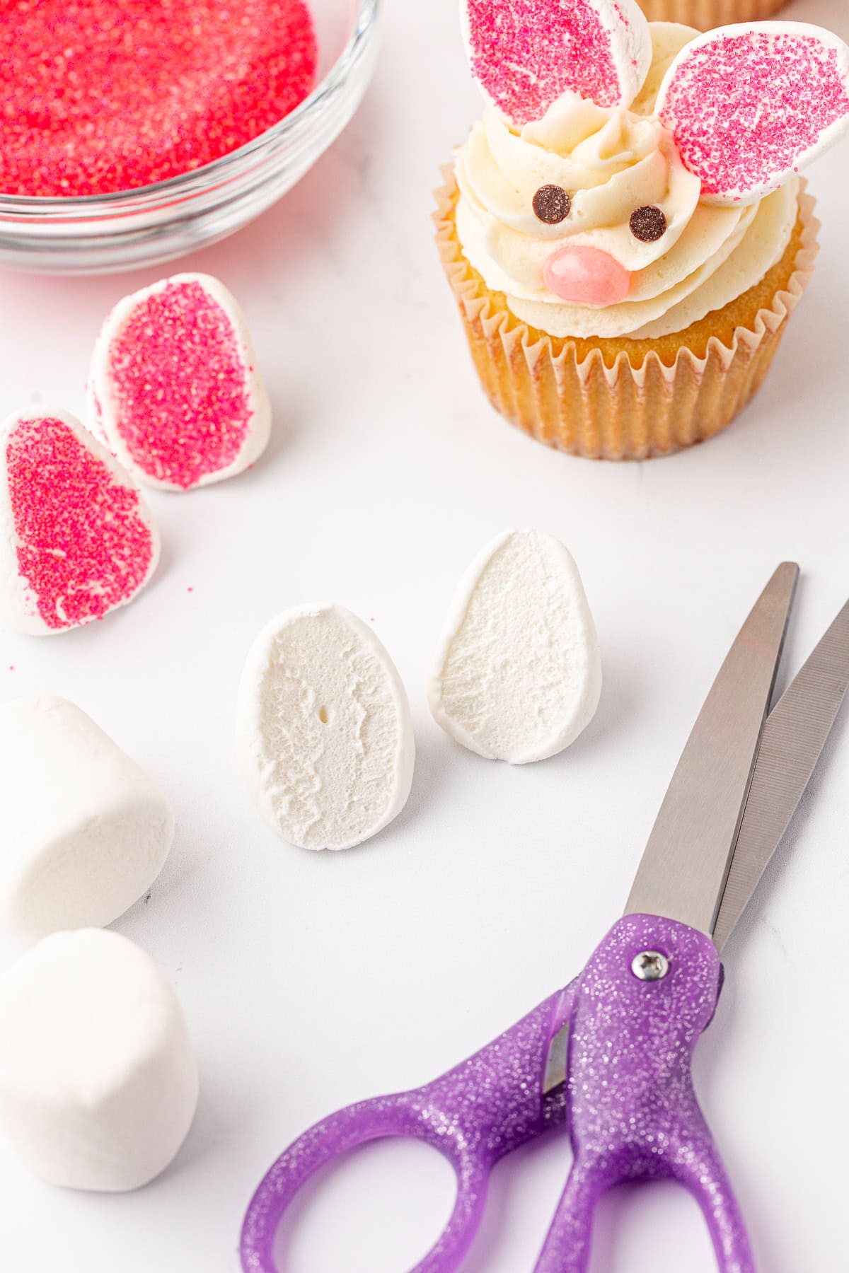 A cupcake decorated as a bunny with pink ears and a face sits near marshmallow halves, pink sugar, a bowl of sprinkles, and purple scissors on a white surface.