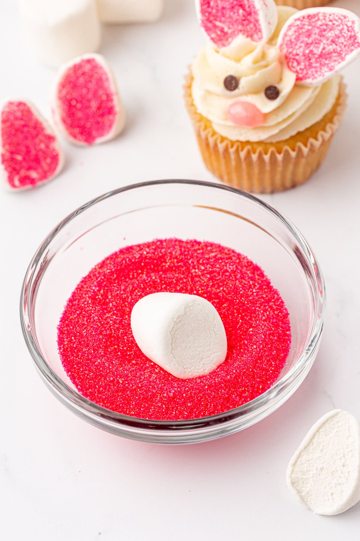 A bowl of pink sugar crystals with a marshmallow being dipped in it, surrounded by marshmallow pieces, and a cupcake decorated like a bunny with frosting, candy decorations, and marshmallow ears.