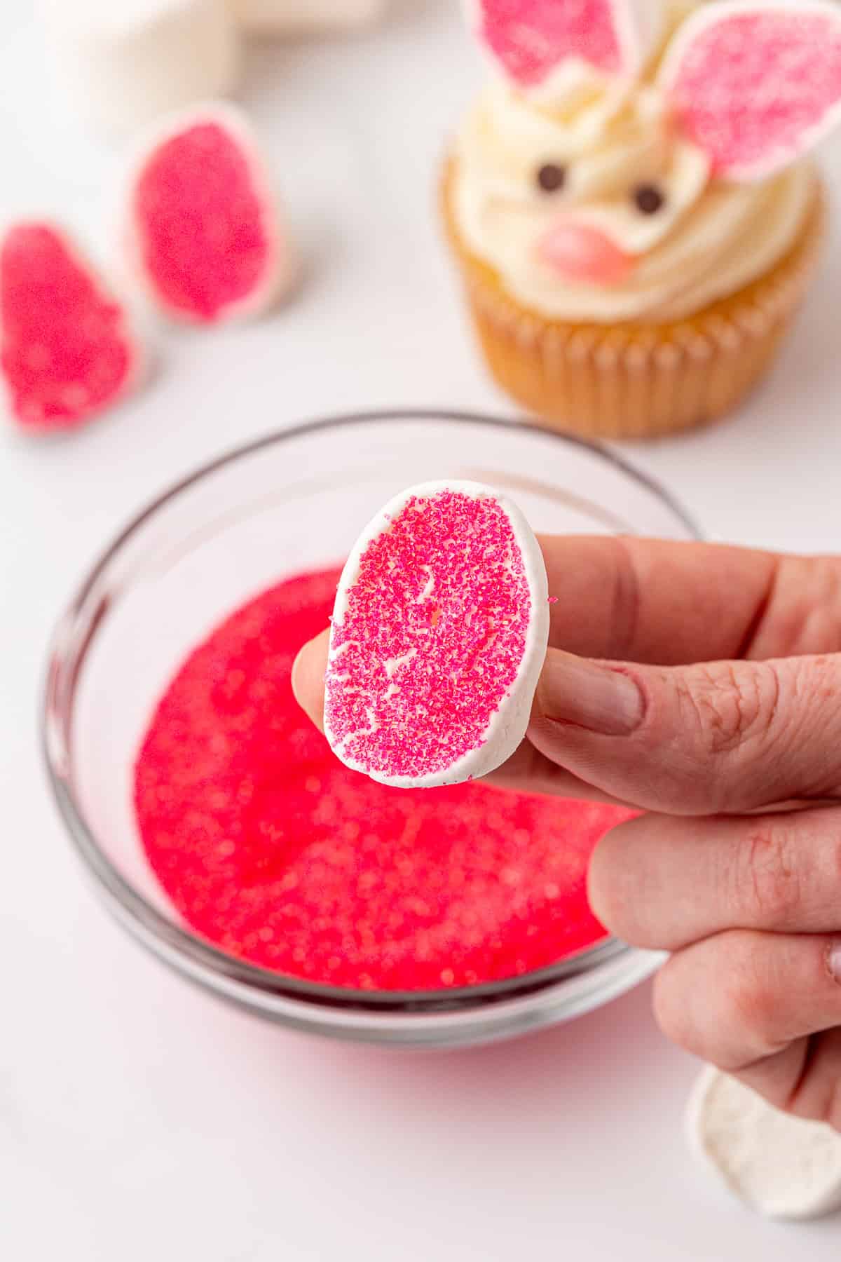 A hand holds a marshmallow half dipped in pink sugar over a bowl of pink sugar, with a bunny-themed cupcake and more pink-dipped marshmallow pieces in the background.