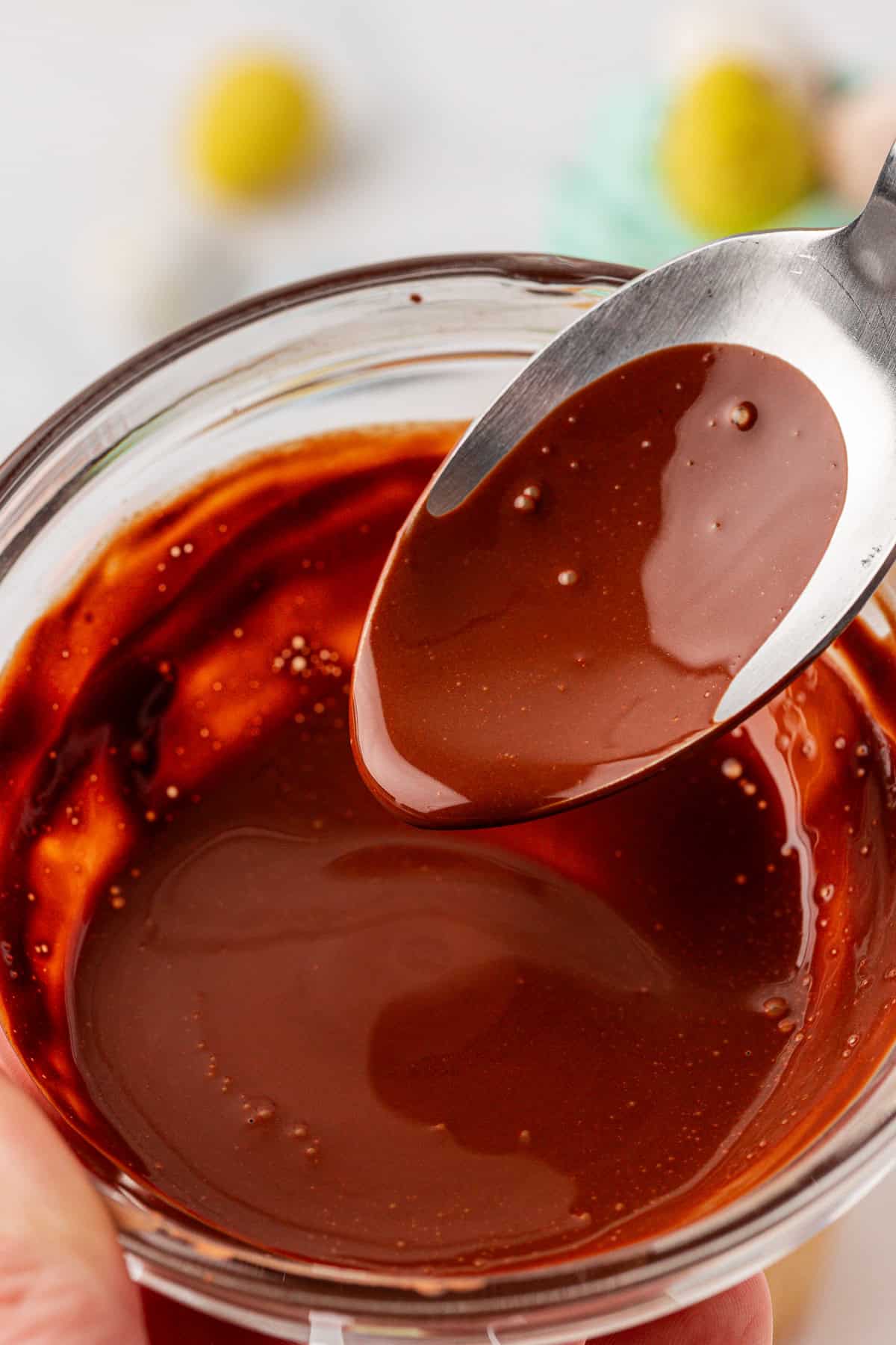 A close-up of a spoon lifting smooth, melted chocolate from a small glass bowl filled with glossy chocolate sauce. The background is softly blurred.