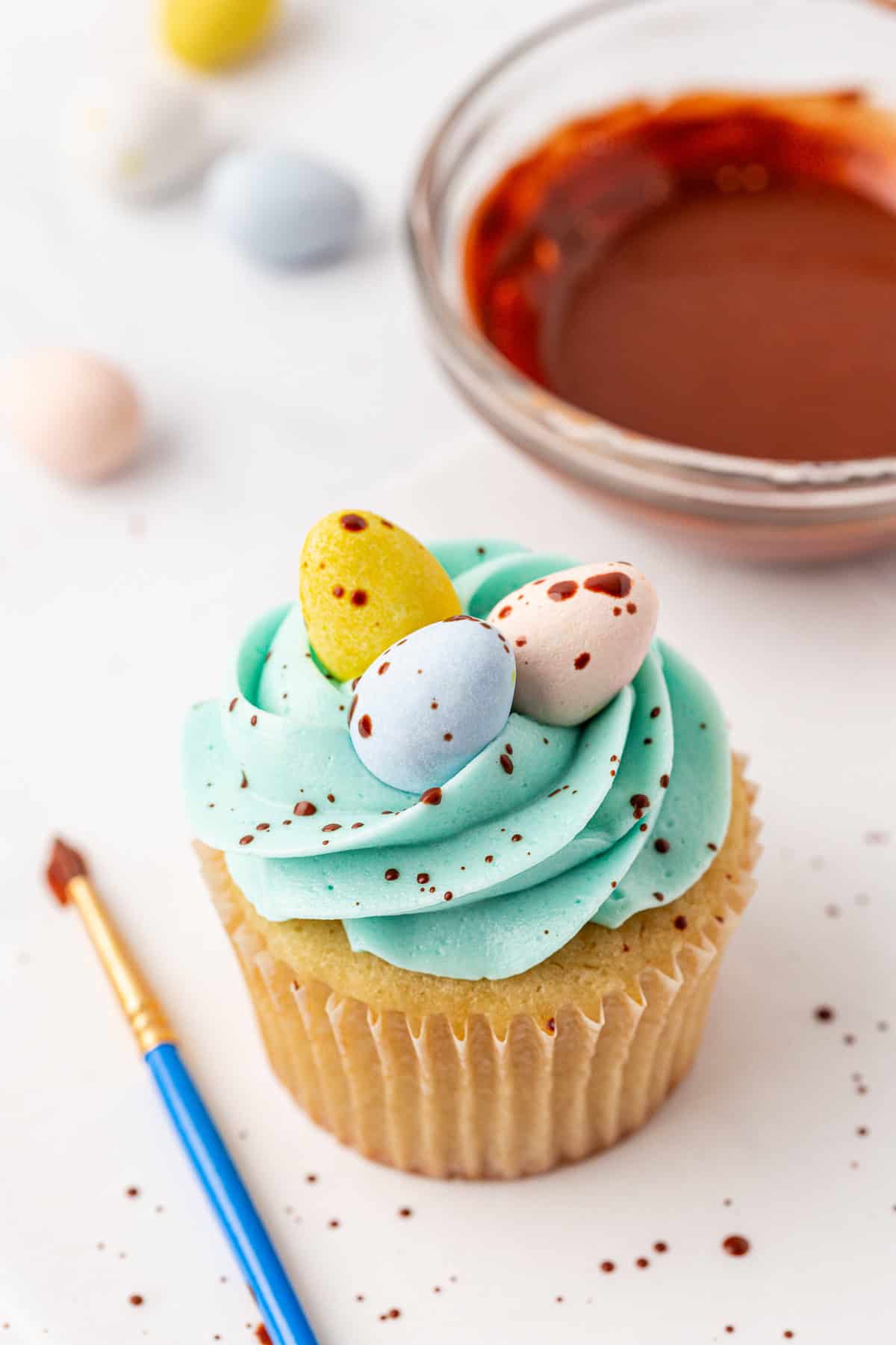 A cupcake with blue frosting is topped with three pastel candy eggs and chocolate speckles. A paintbrush and a bowl of melted chocolate sit nearby on a white surface.
