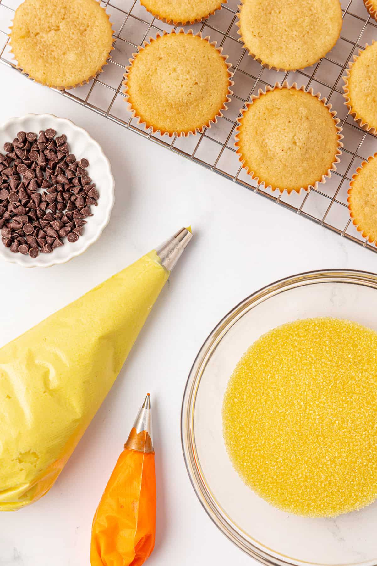 Unfrosted cupcakes on a cooling rack, a bowl of chocolate chips, yellow and orange frosting in piping bags, and a bowl of yellow sugar sprinkles on a white surface.