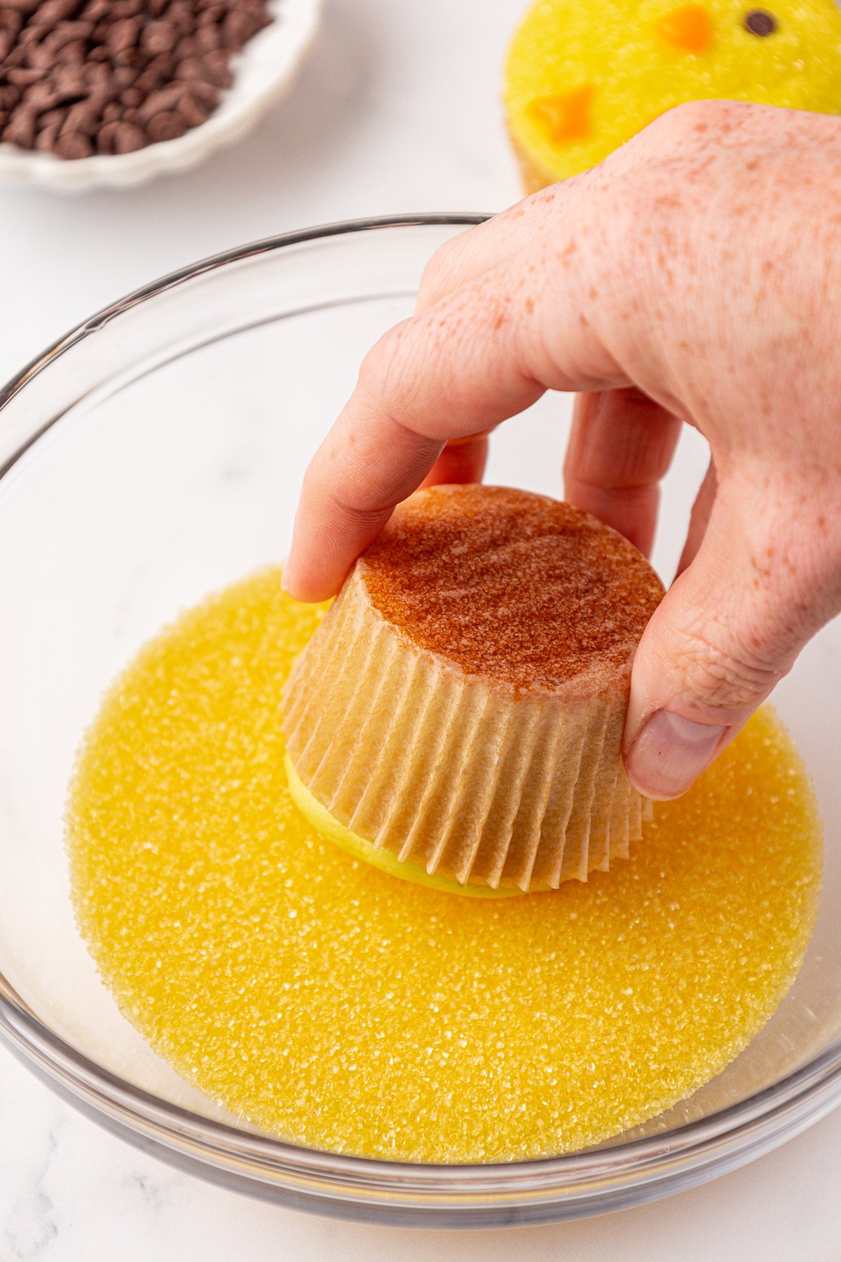 A hand dips a cupcake with a brown paper liner into a bowl of yellow sugar crystals. Chocolate chips and a decorated cupcake are visible in the background.