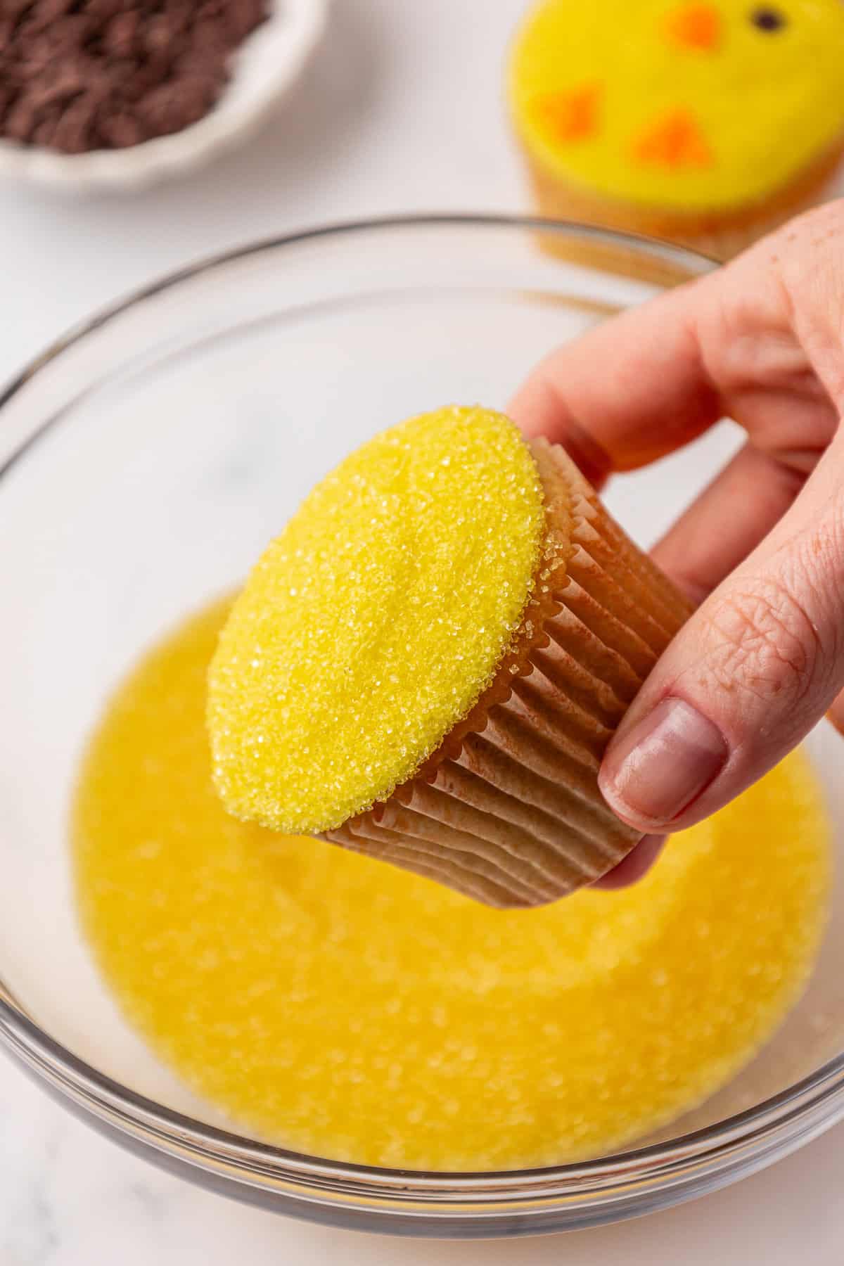 A hand holds a cupcake with yellow sugar sprinkles over a bowl containing more yellow sugar. Another decorated cupcake and a bowl of chocolate chips are visible in the background.