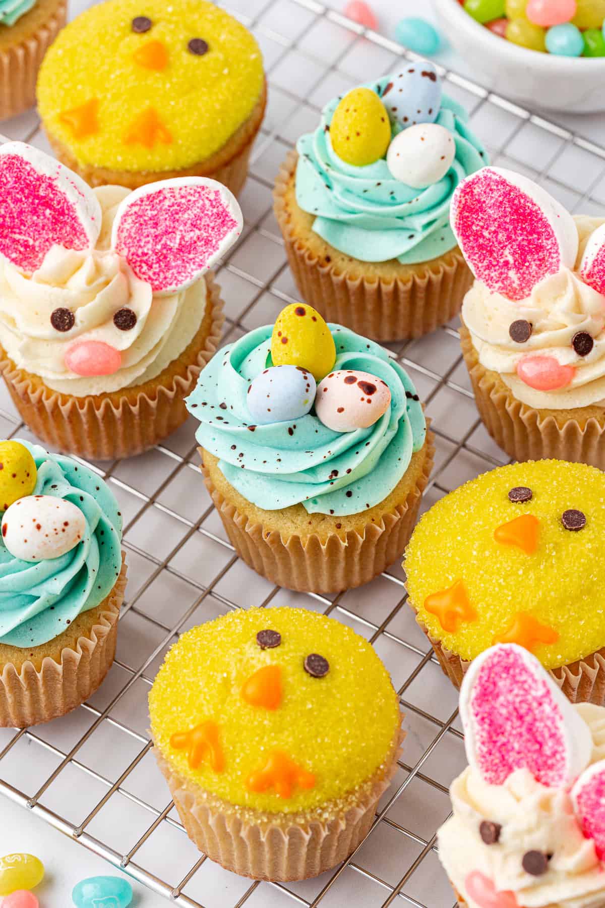 A variety of Easter-themed cupcakes on a cooling rack, decorated as yellow chicks, bunny faces with pink ears, and blue swirled nests topped with candy eggs.