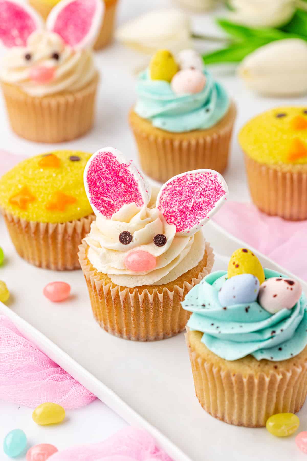 A platter of decorated cupcakes, including one with bunny ears and face made of frosting, and others topped with pastel-colored eggs and yellow sprinkles. Jellybeans and flowers are scattered around for a festive spring theme.